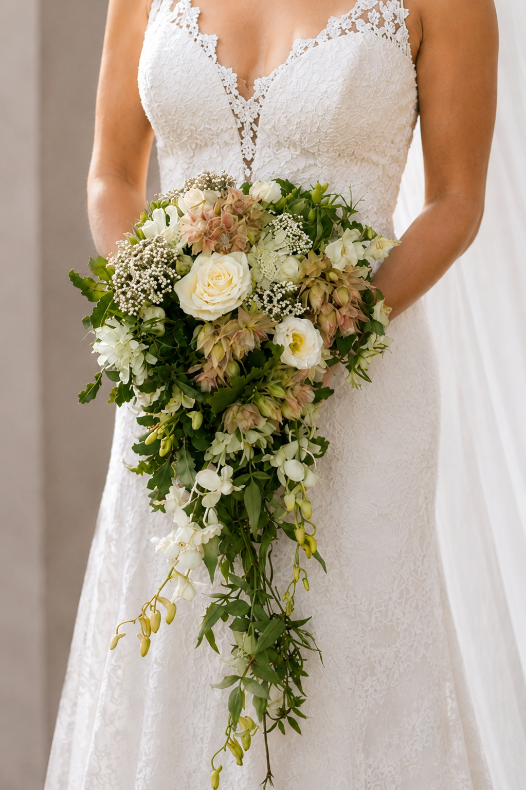 A bride in a white lace wedding dress holding a cascading or shower bouquet of seasonal romantic flowers. Jasmine, orchids, freesia.  The dress features lace detailing around the neckline.
