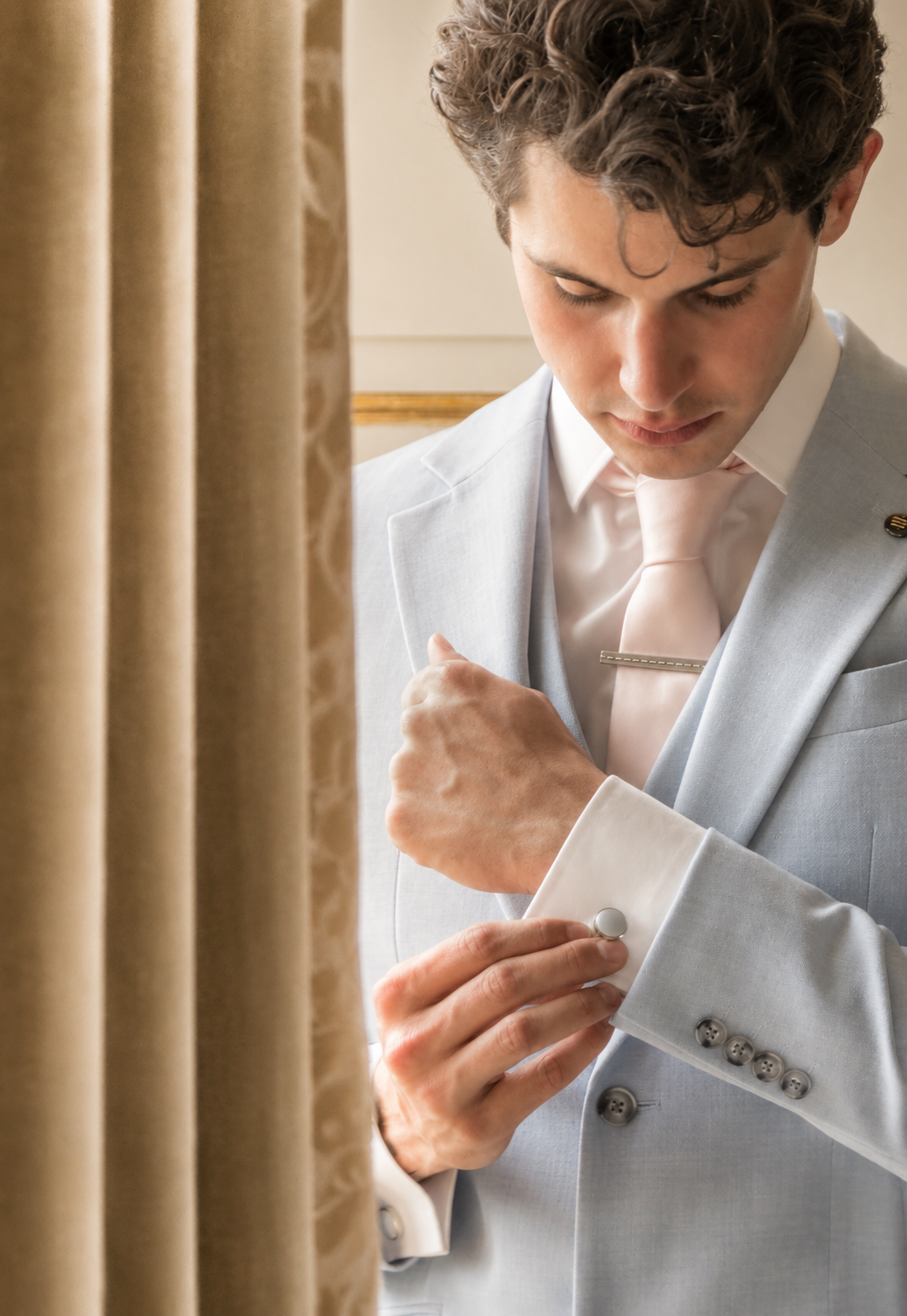 A man in a light gray suit adjusting his cufflinks while looking down, behind a beige curtain.