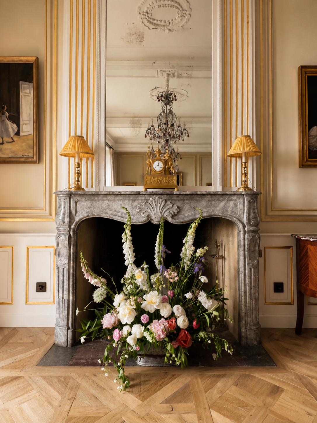 Elegant living room with a marble fireplace, floral arrangement with white, pink, and purple flowers, large mirror, gold table lamps, and classic artwork.