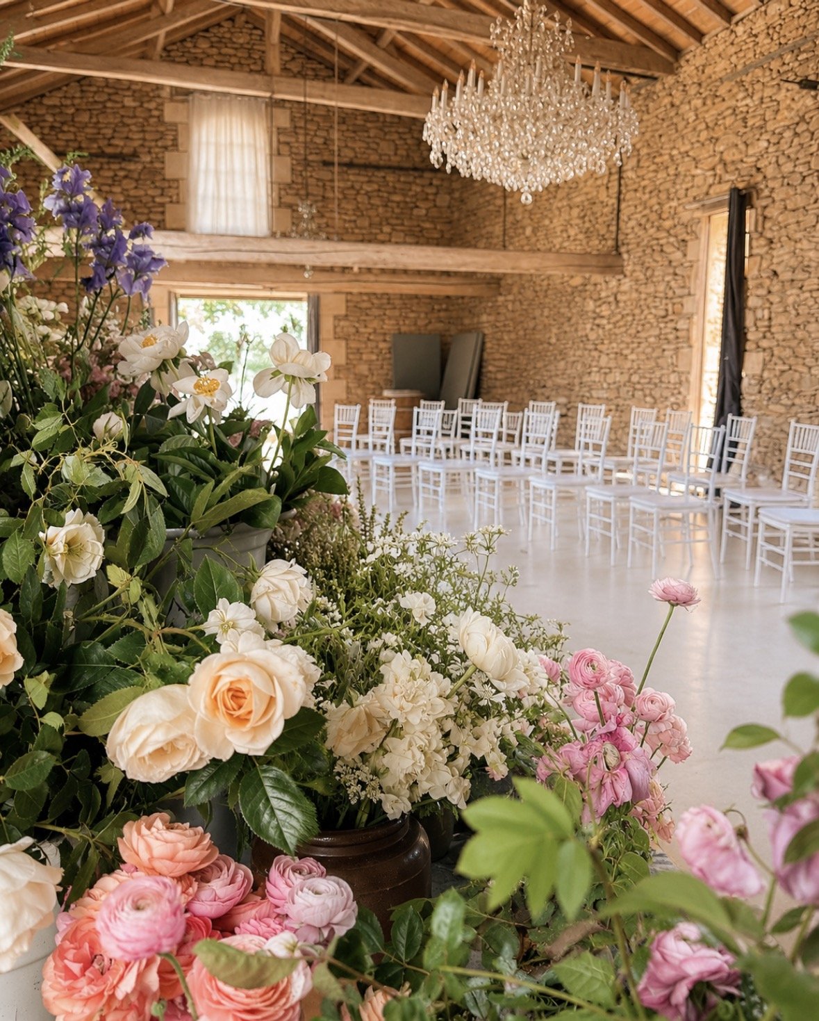 Flower arrangement with white, pink, and purple flowers in a rustic setting with white chairs arranged in rows, stone walls, and a chandelier hanging from the ceiling.