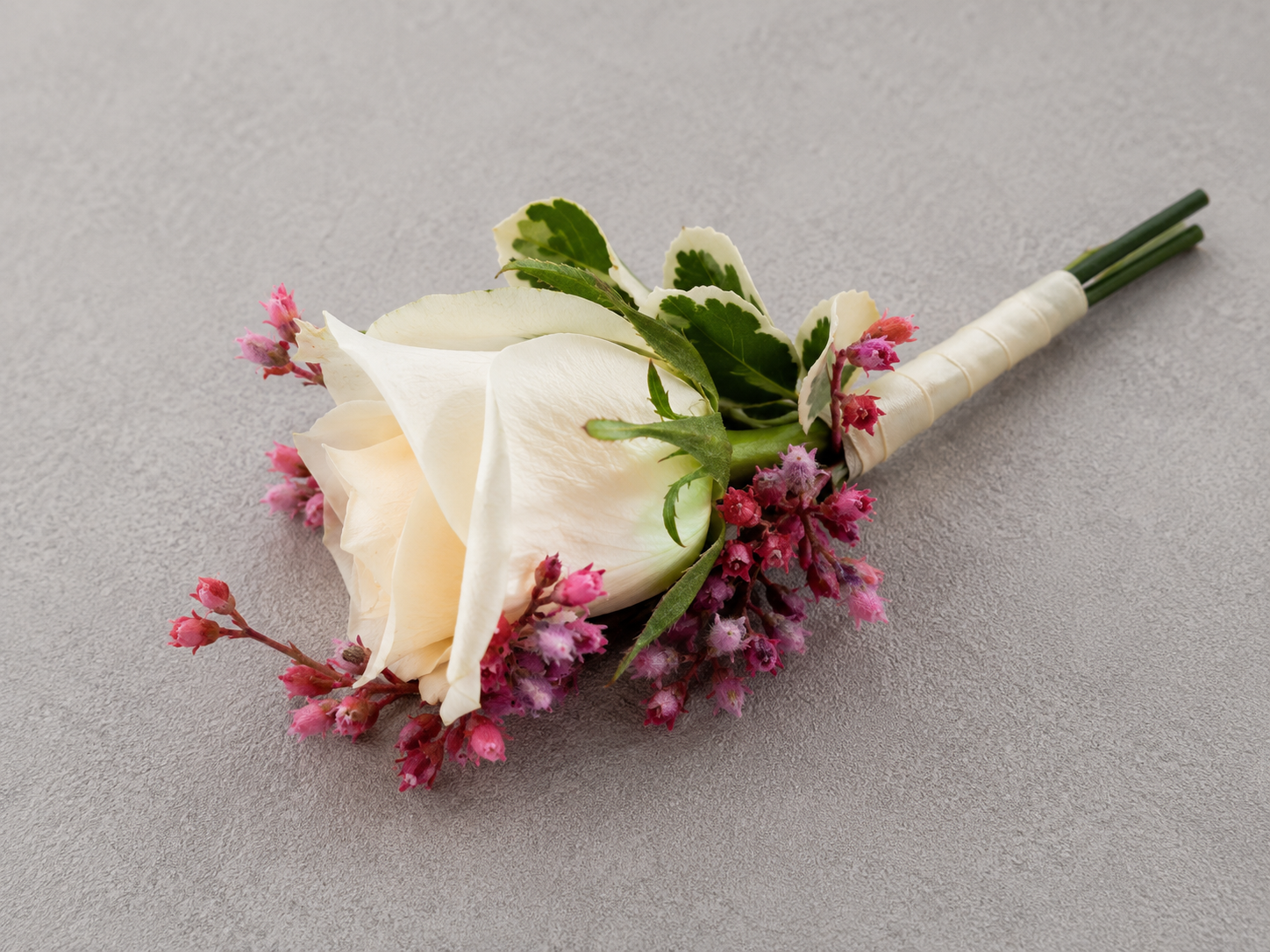 Floral boutonniere with white rose, pink accent flowers, and variegated green leaves, wrapped with white ribbon on grey background.