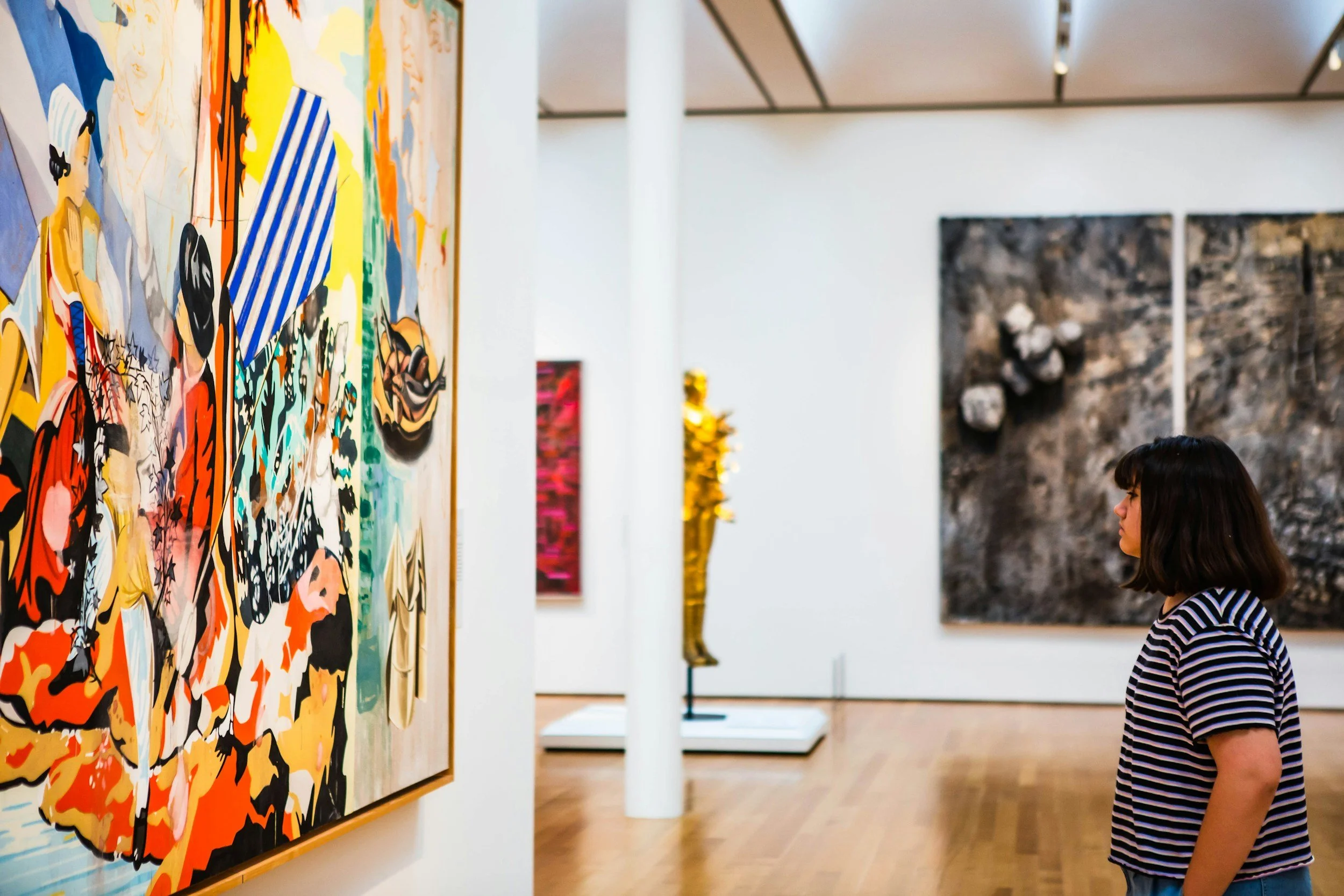 A woman with dark hair and a striped shirt is observing colorful abstract paintings in an art gallery. The paintings are vibrant and expressive, hung on white walls with a wooden floor beneath.