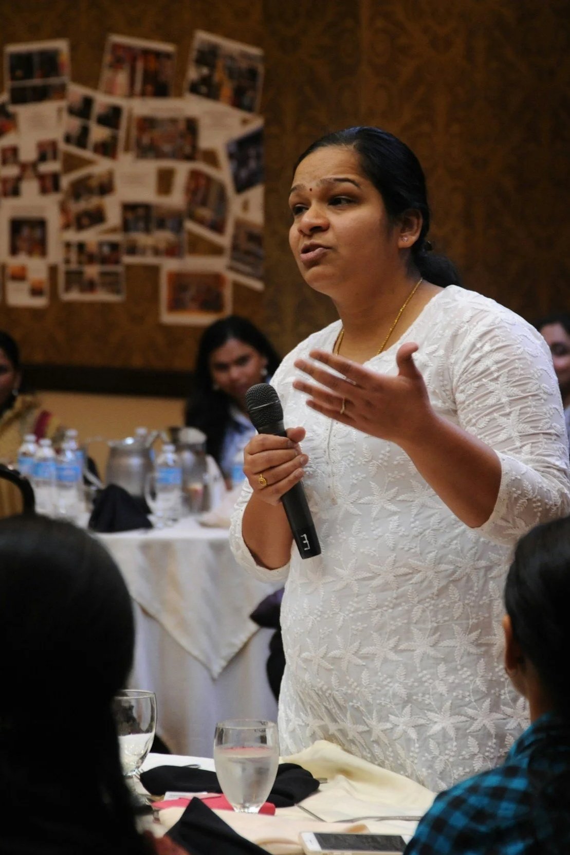 A woman in a white embroidered kurta speaking into a microphone at a formal event, with people seated at tables in the background.