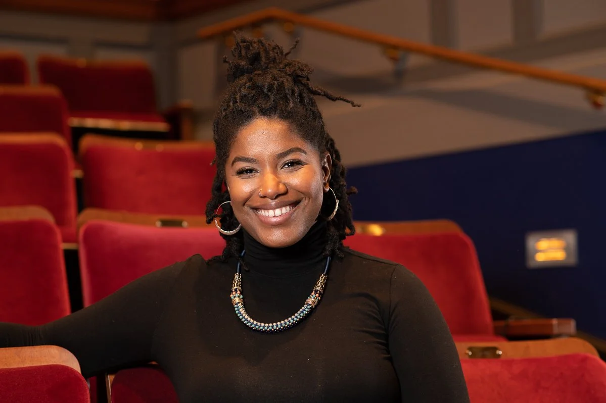 A woman with dark skin and dreadlocked hair styled in an updo, smiling while sitting in an auditorium with red seats and a blue wall background, wearing a black turtleneck, hoop earrings, and a beaded necklace.