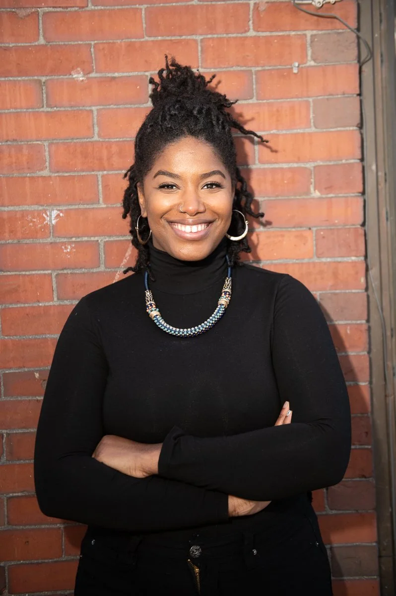 A woman with dark skin and dreadlocked hair styled in a bun, smiling with crossed arms against a brick wall background, wearing a black turtleneck, hoop earrings, and a beaded necklace.
