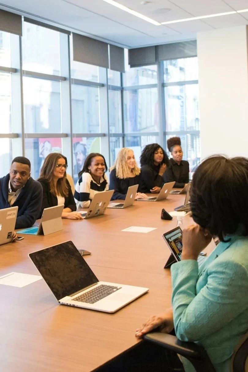A diverse group of six people sitting at a conference table with laptops, engaged in a meeting in a modern office with large windows.