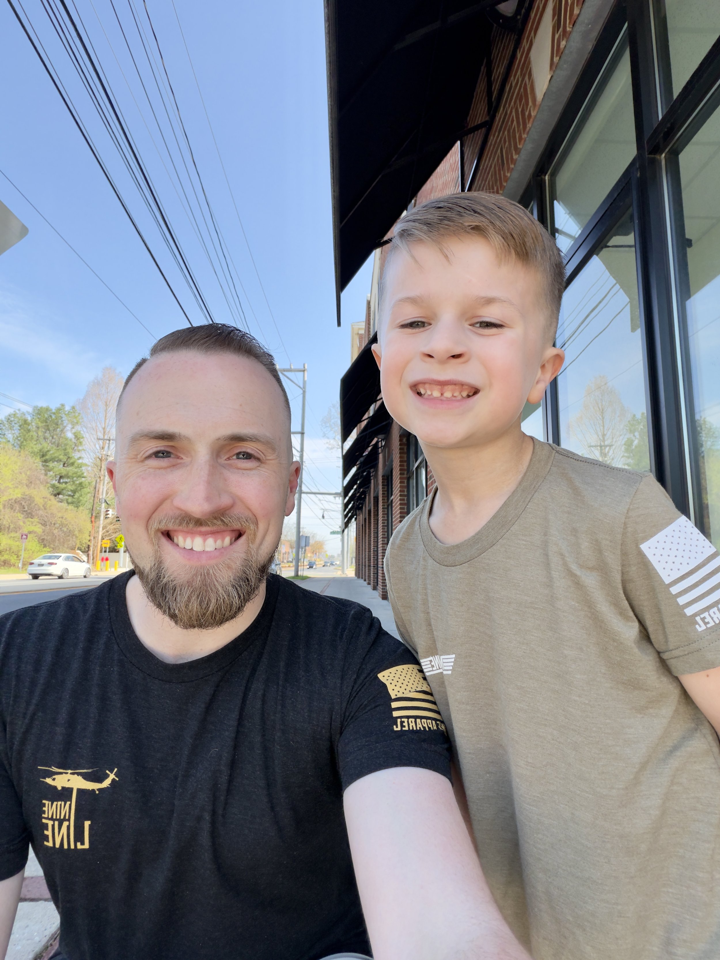 A smiling man with a beard and a young boy with short hair taking a selfie outdoors on a sunny day, with a building with large windows and a blue sky in the background.