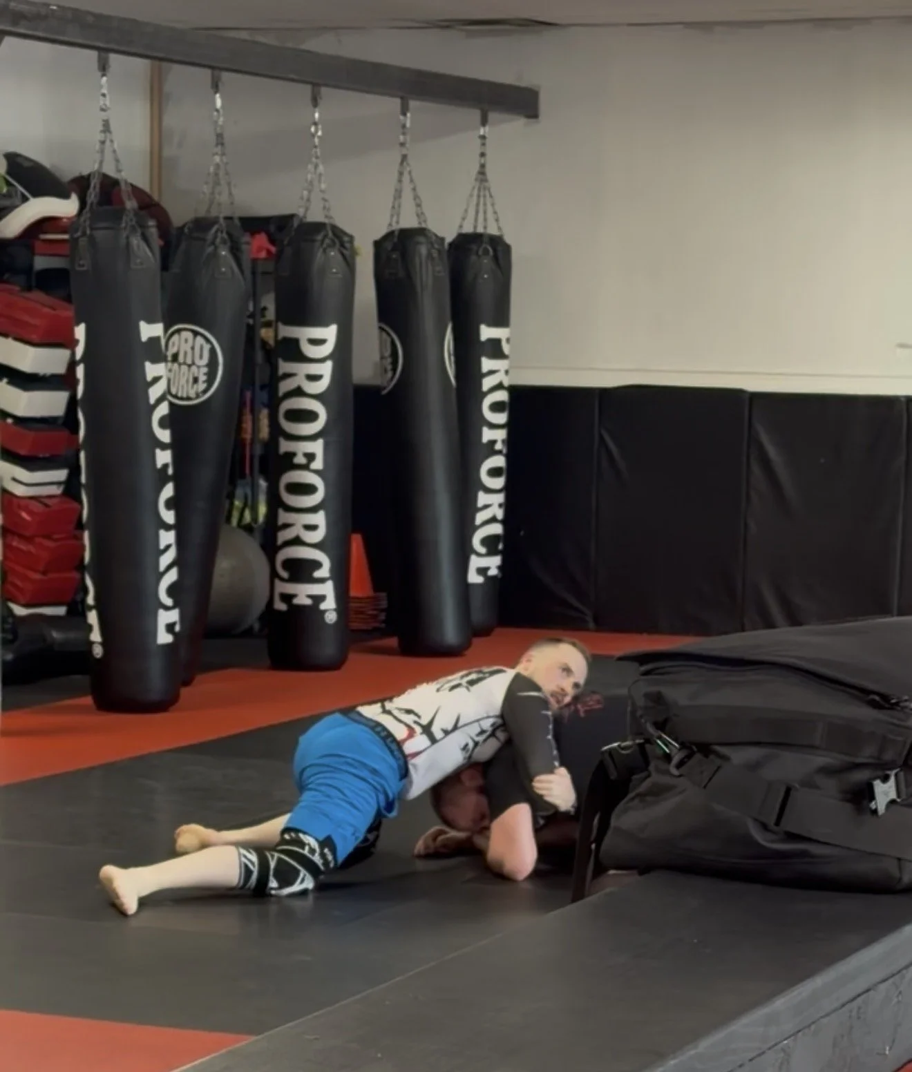 Two people practicing Brazilian Jiu-Jitsu on a padded training mat in a gym, with punching bags in the background.