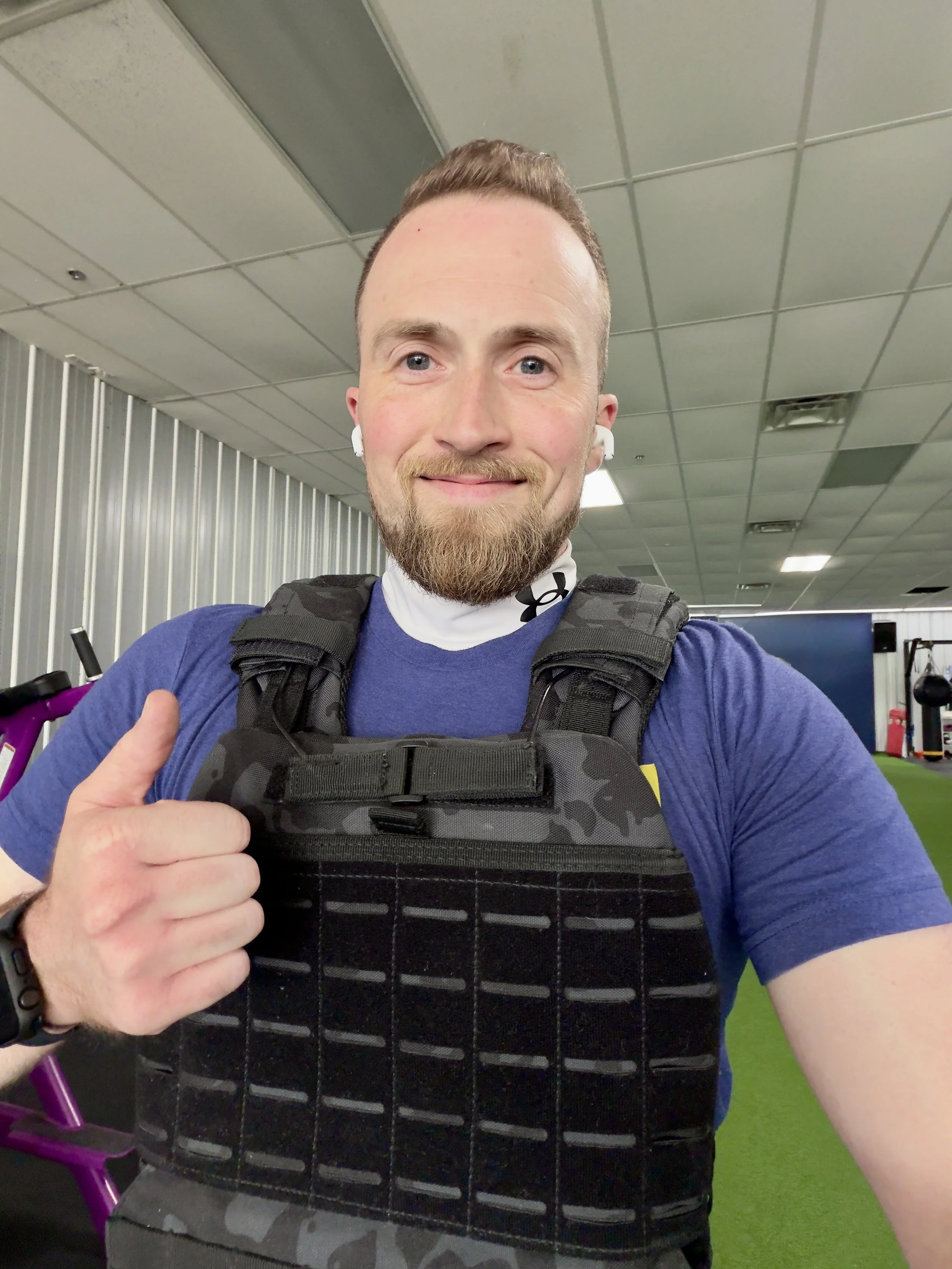Close-up of a smiling man at the gym, wearing a navy blue shirt, a black workout vest, wireless earbuds, and a black wristband, showing a thumbs-up.