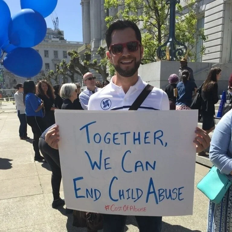 Ryan holds a sign that reads, "Together, We Can End Child Abuse," during a public outdoor event. There are other people and balloons in the background.