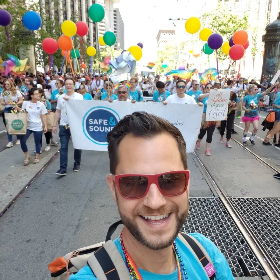 Ryan taking a selfie at a pride parade with a large crowd of people holding rainbow flags, balloons, signs, and wearing colorful clothing in the background.