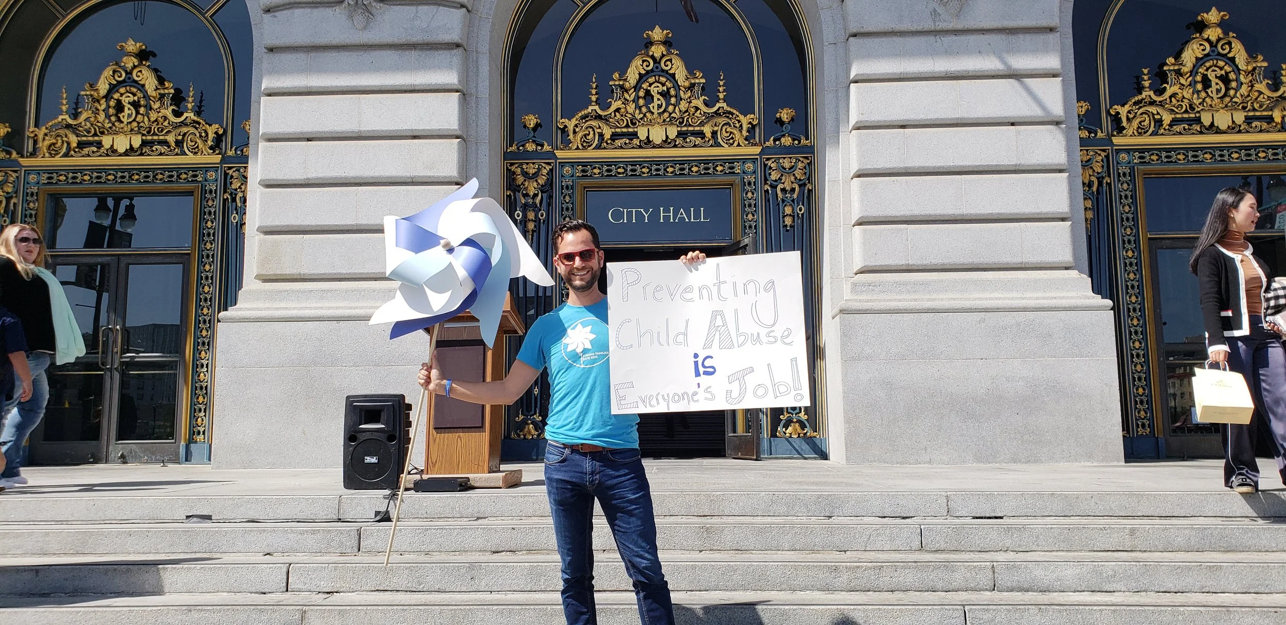 standing on the steps of a city hall holding a sign that reads 'Preventing Child Abuse is Everyone's JOB!' next to a large blue and white pinwheel sculpture. He is smiling and wearing a blue t-shirt and sunglasses, with other people walking by in the background.