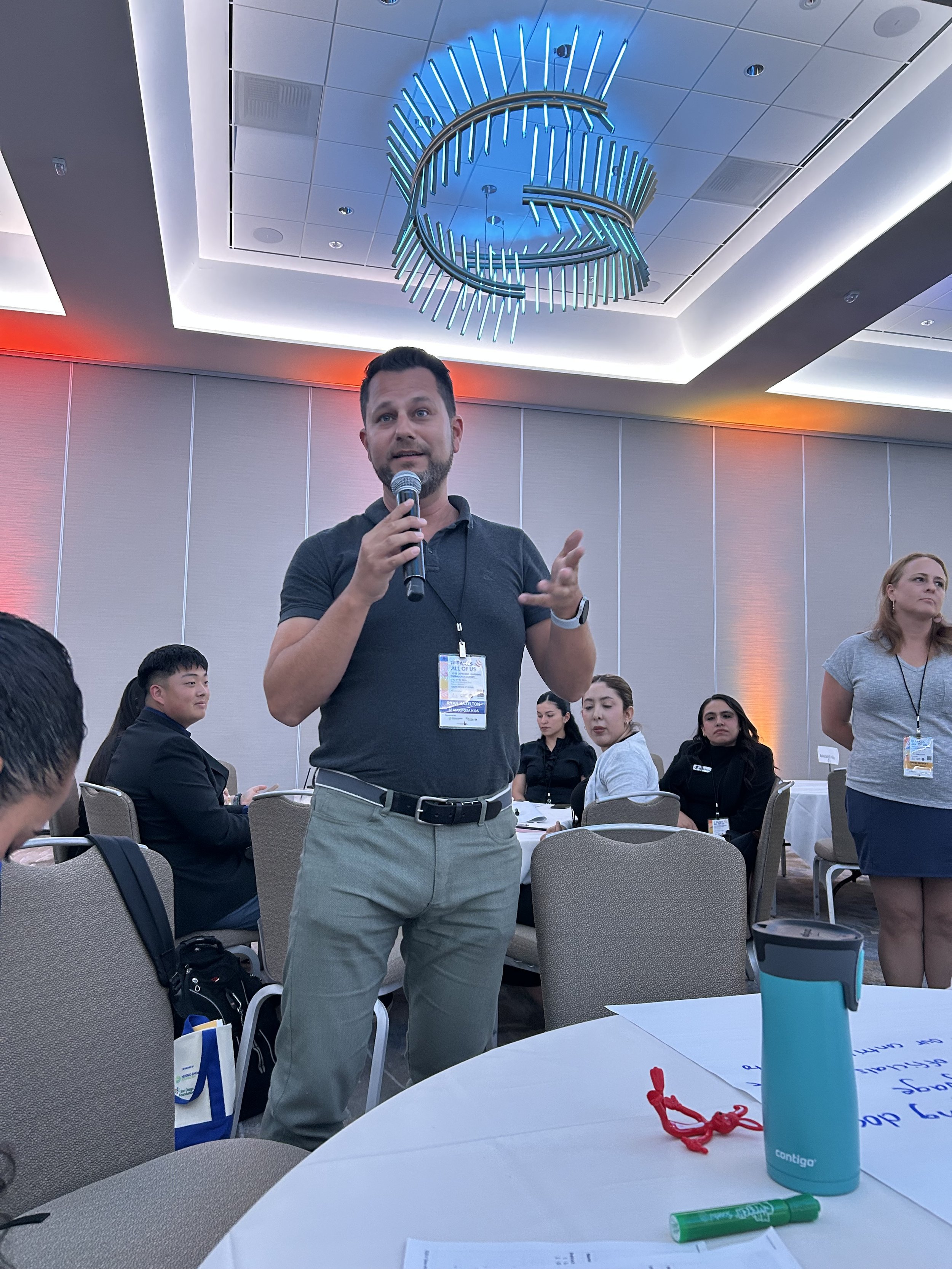Ryan is speaking into a microphone at a conference with several people seated around him in a brightly lit room.