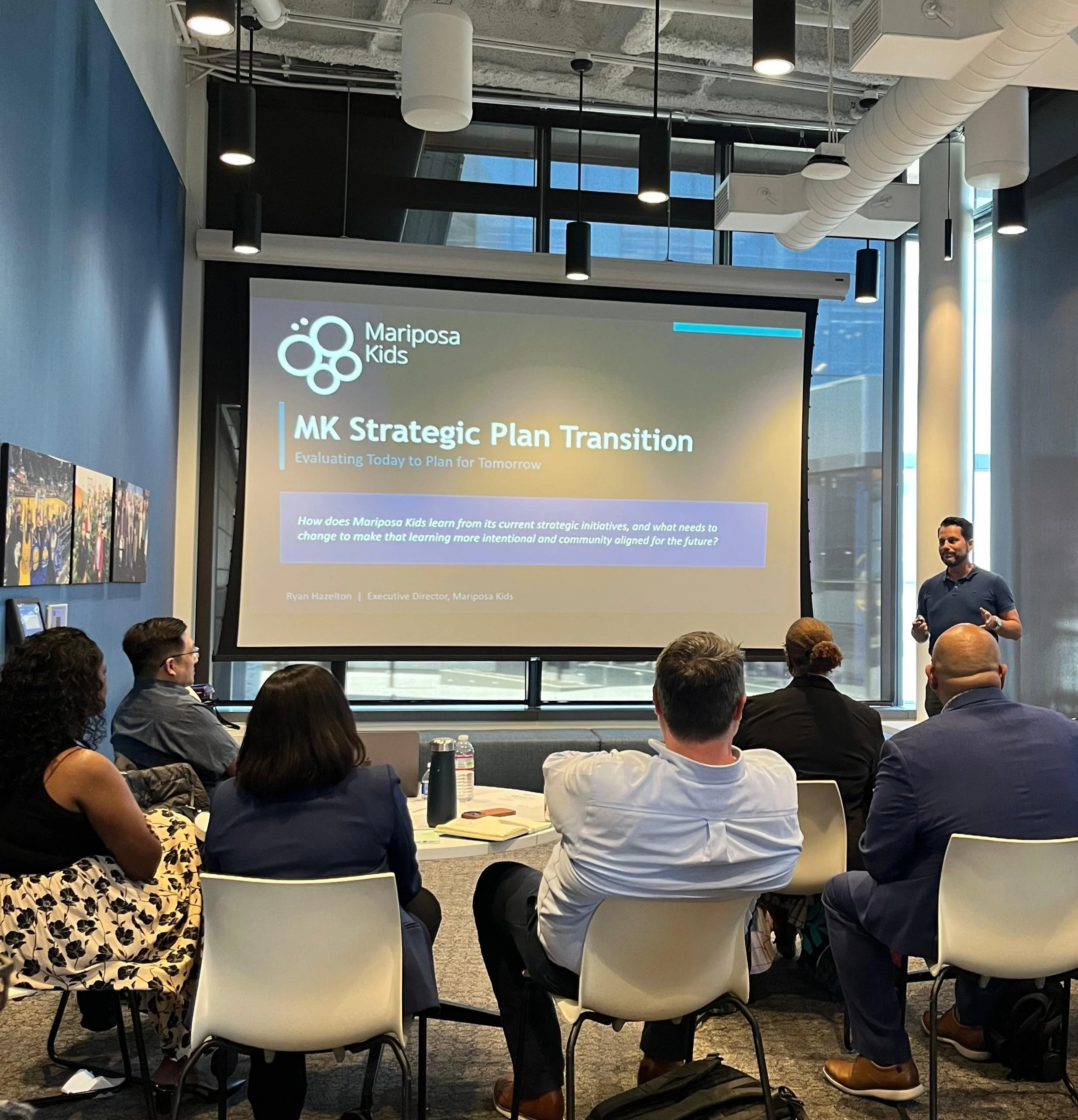 A group of people attending a presentation in a conference room. The speaker, a man, is standing near a screen displaying a slide titled 'Mariposa Kids,' with additional text and a logo. The room has modern design elements, including large windows, pendant lights, and visible air ducts.