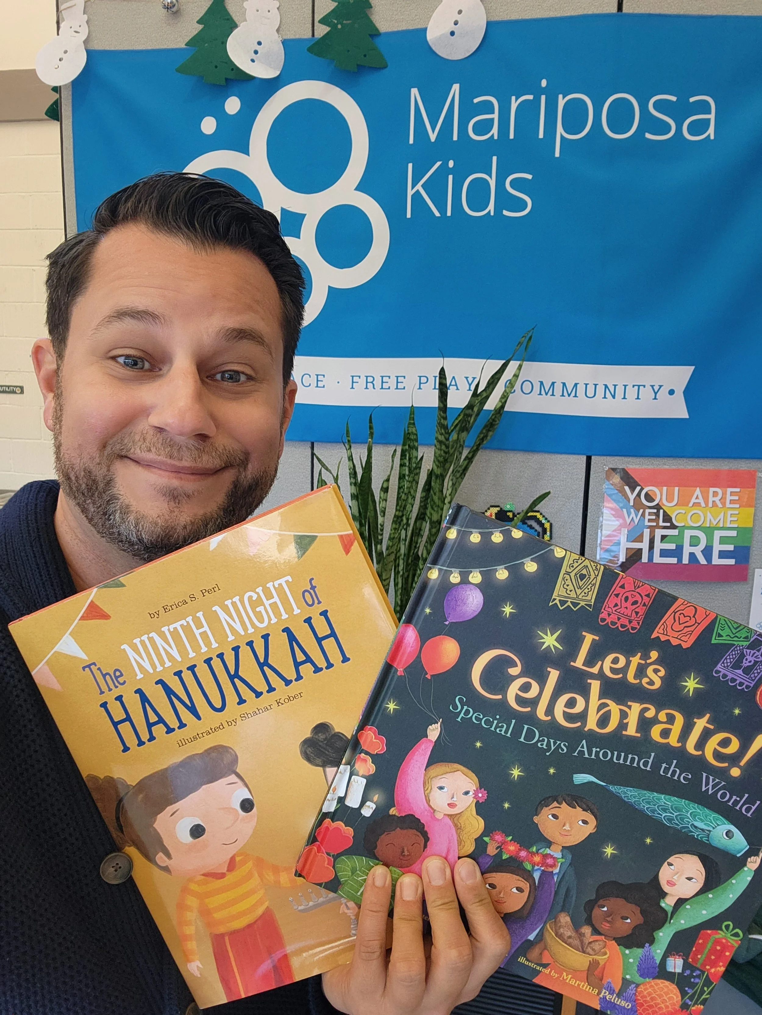 Ryan taking a selfie with a background of a blue Mariposa Kids poster decorated with paper snowmen and Christmas trees. He is holding two colorful books about Hanukkah and celebrating special days around the world.