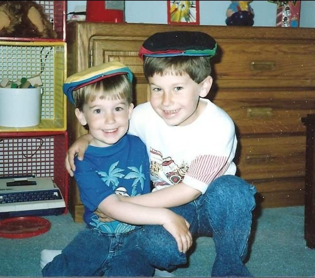 Two young boys sitting on a carpeted floor, smiling at the camera, with small stackable travel pillows on their heads. The boy on the left is wearing a blue shirt with a palm tree design, and the boy on the right is wearing a white t-shirt with red and black graphics. Behind them, there is a wooden dresser and a colorful toy storage unit.