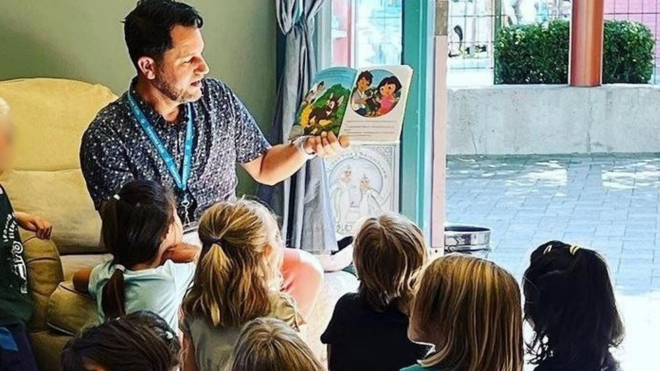 Ryan reading a children's book to a group of children sitting on the floor indoors near a large window.