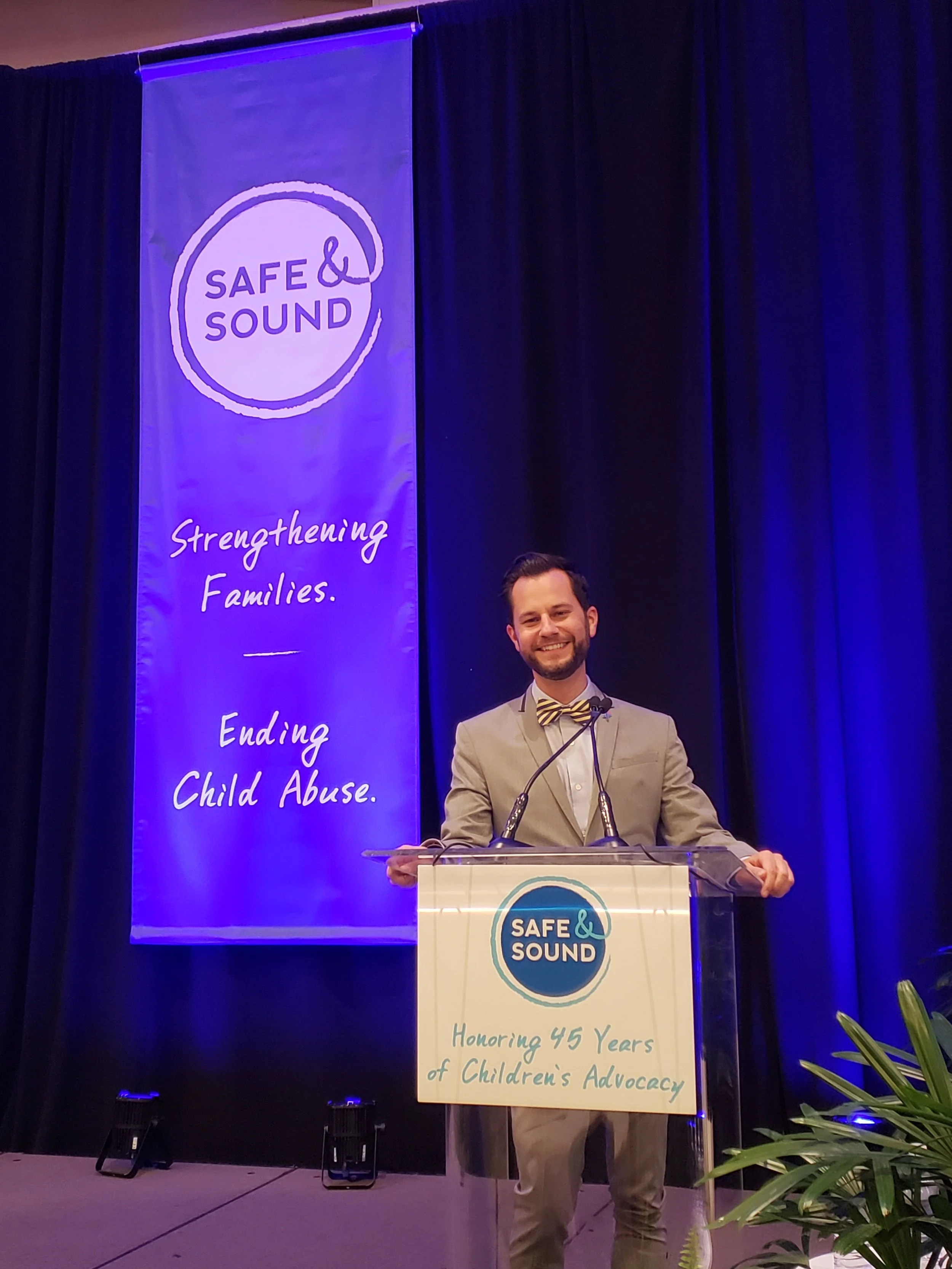 Ryan standing at a podium with a sign that reads "Honoring 45 Years of Children's Advocacy" at an event for Safe & Sound, with a large blue banner behind him displaying the Safe & Sound logo and slogan to end child abuse.