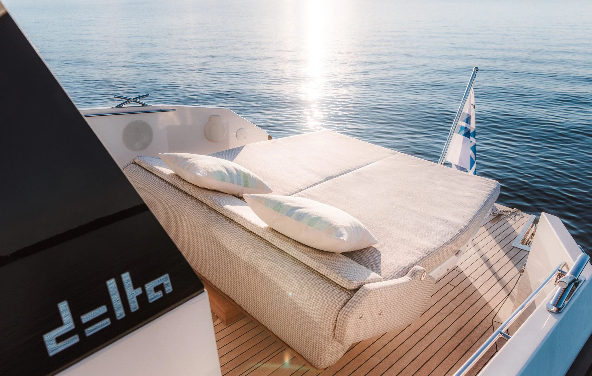 Sunlit view of the deck of a boat with a lounge chair, pillows, and a small flag, overlooking calm water.