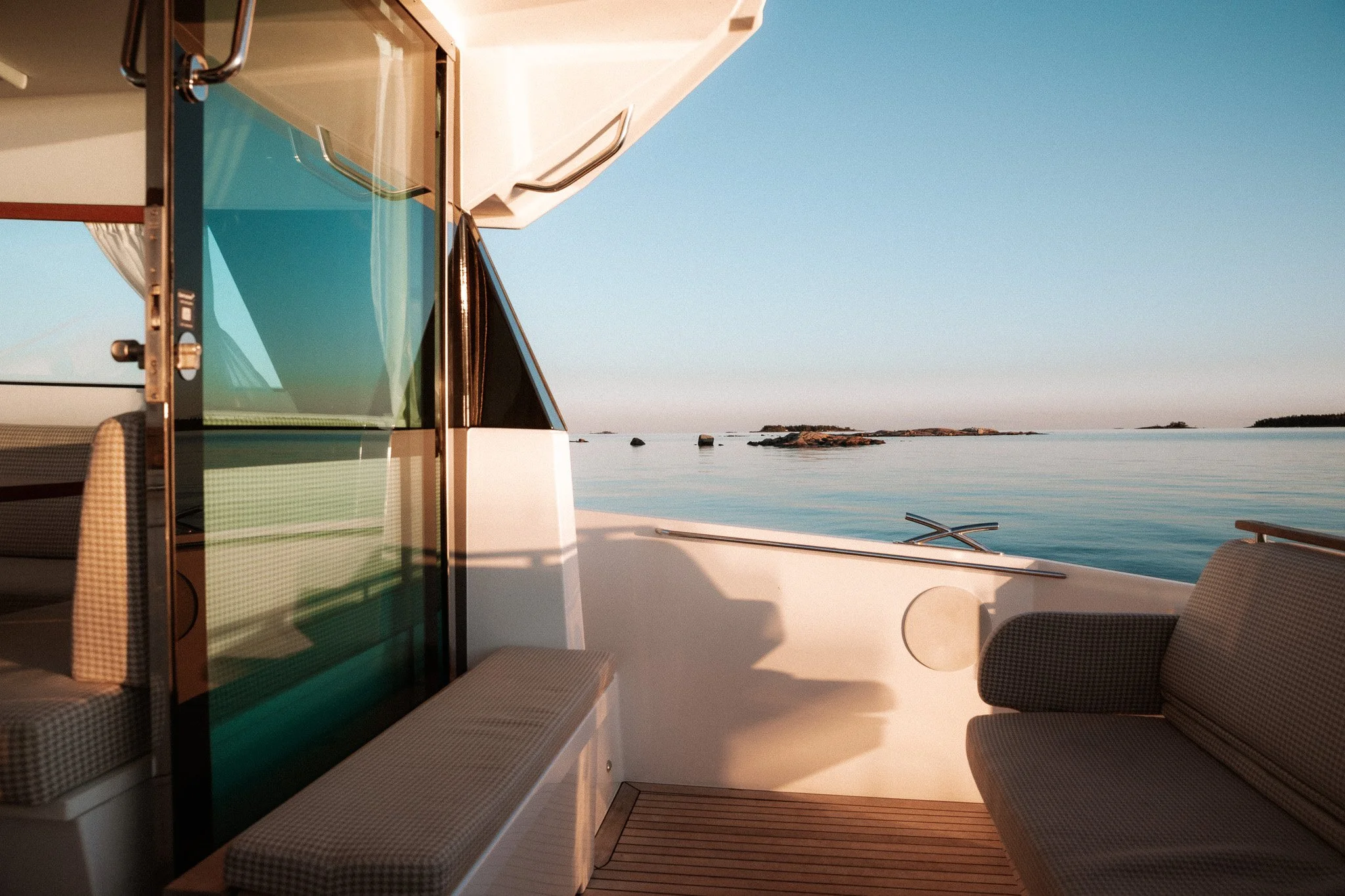 View from the deck of a boat showing part of the cabin with glass doors, outdoor seating with checkered cushions, and a calm body of water with rocks and small islands in the distance under a clear sky.