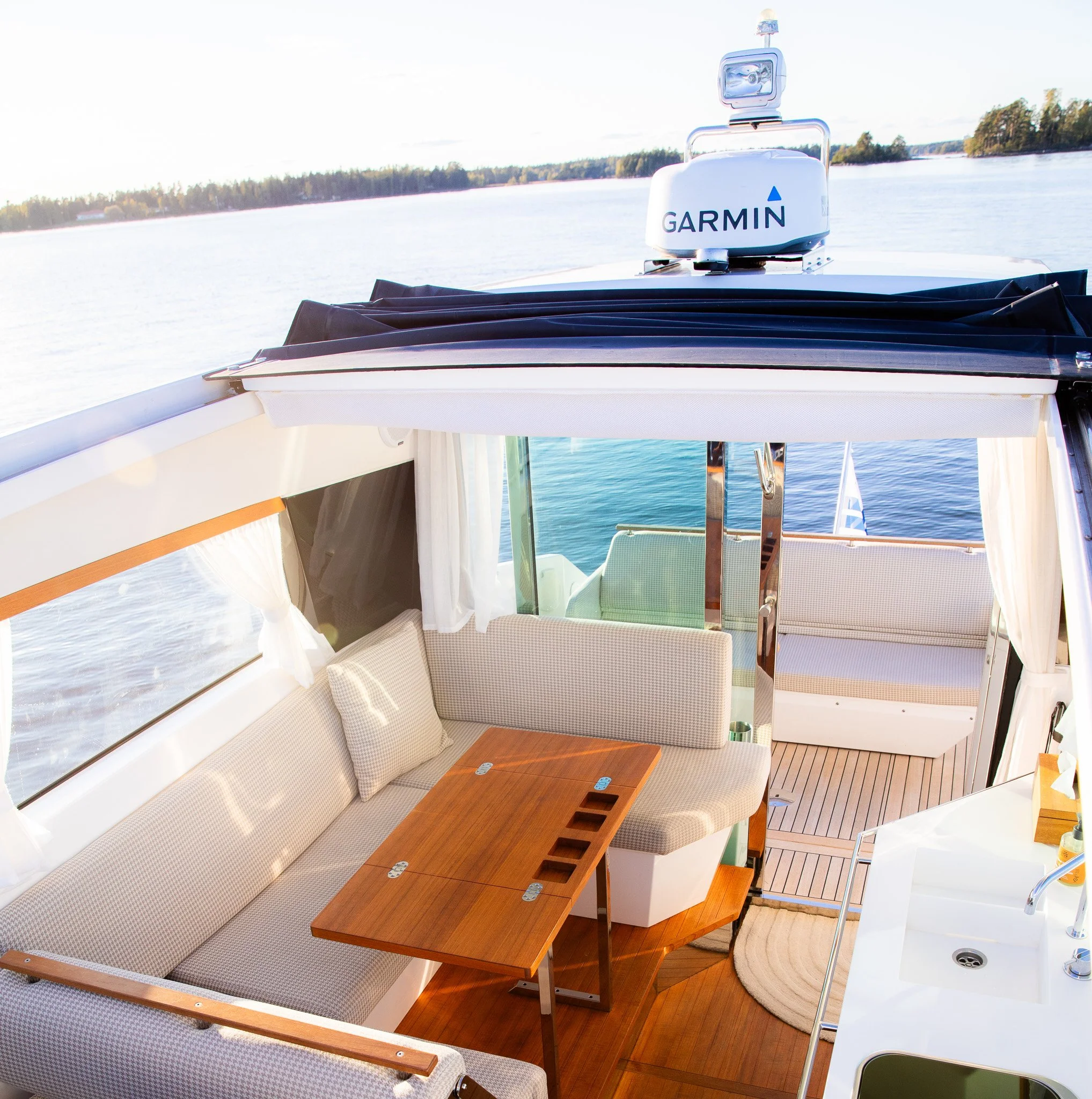 View from inside a boat showing a white boat cabin with beige seating, a small wooden table, and a white Garmin radar on top of the cabin, overlooking a body of water with distant trees.