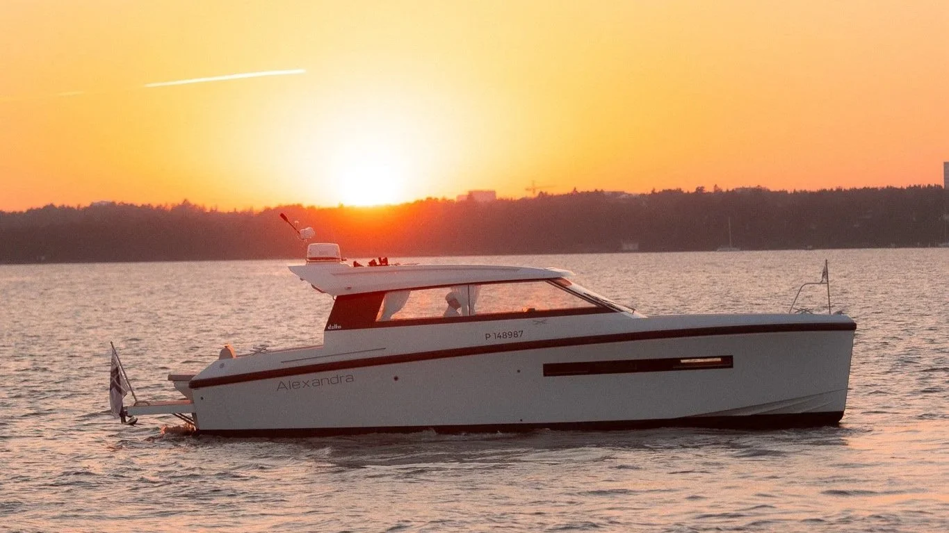 A white yacht named Alexandra sailing on calm water during a sunset with an orange sky and distant land on the horizon.