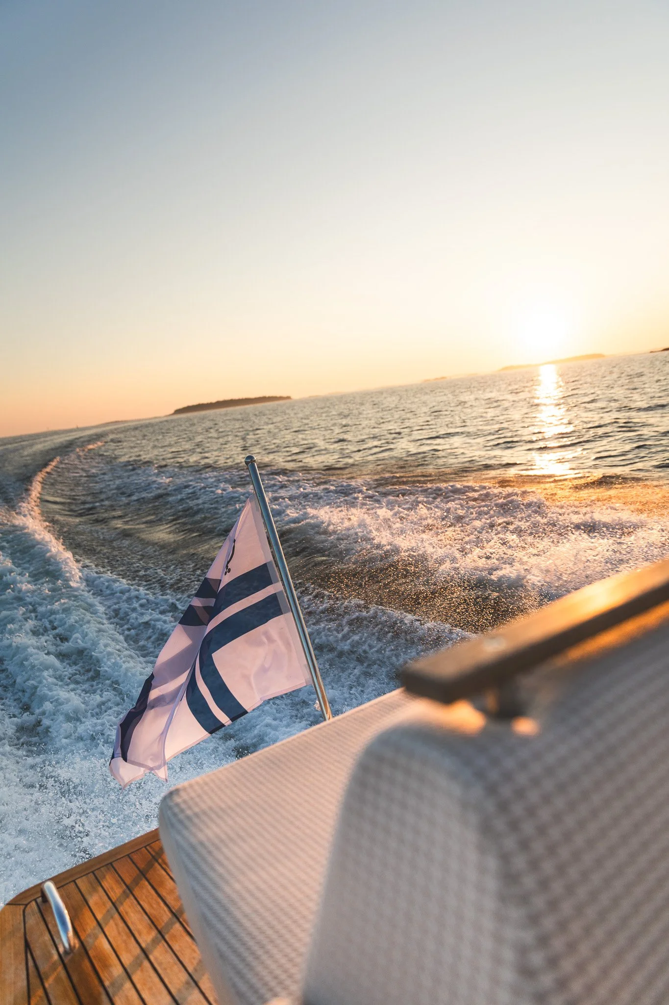 View from a boat at sunset, showing the boat's wake on the water, a flag with a blue and white design, and an island in the distance.