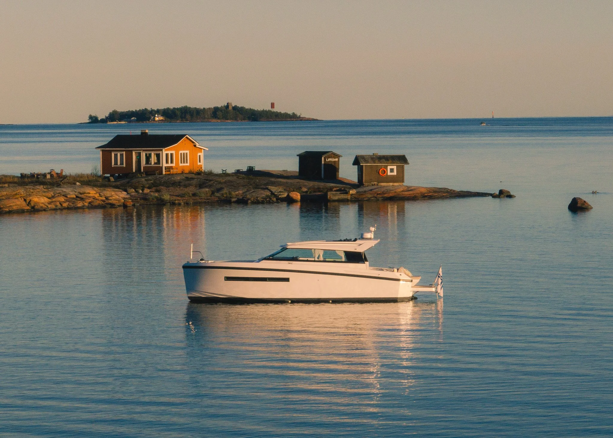A white boat floating on calm water near a rocky shoreline with small houses, a small island with trees and structures, and a distant lighthouse in the background during sunset.