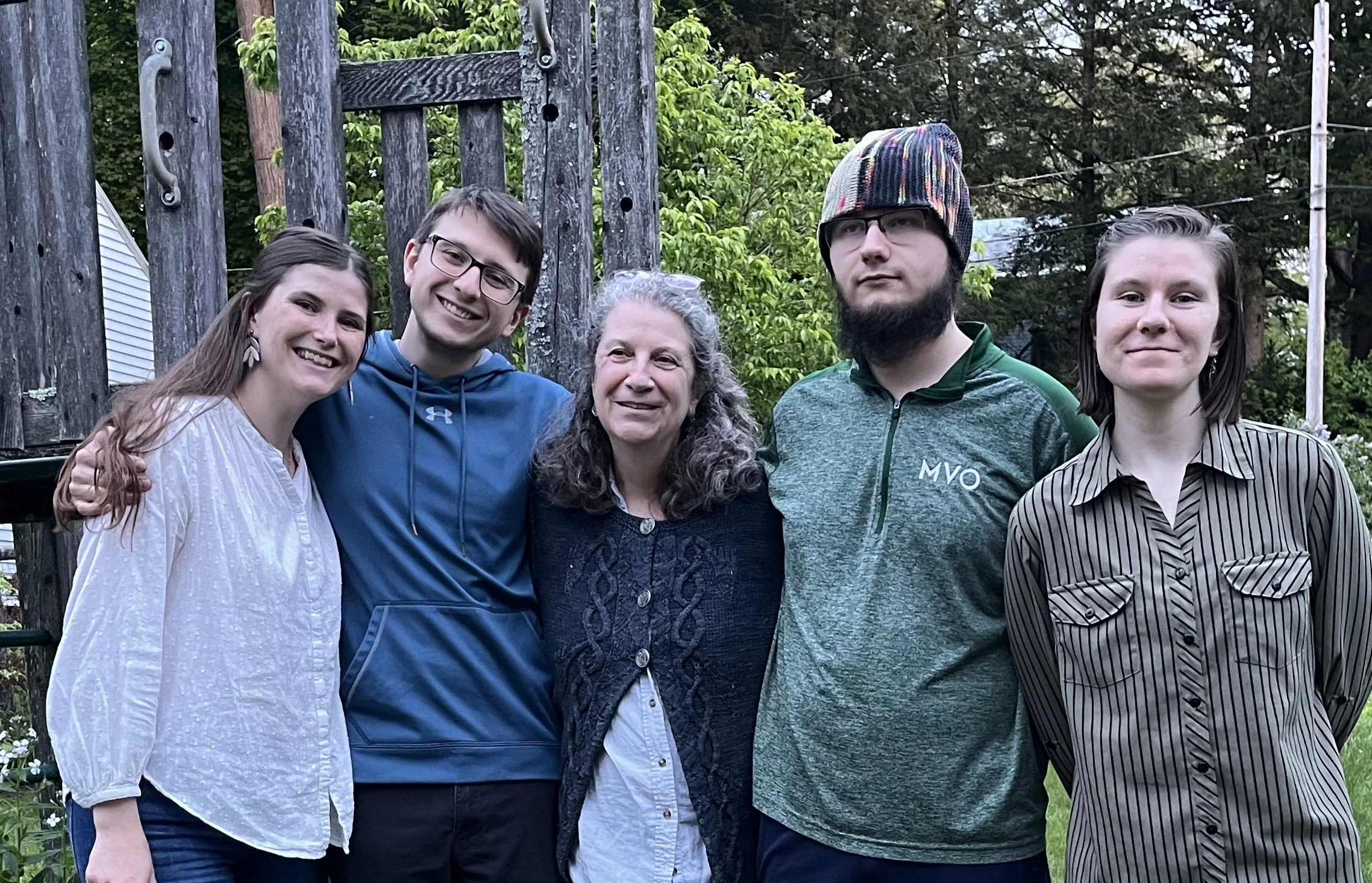 Group of five people standing outdoors in a garden, smiling at the camera. The group includes three women and two men, with a wooden structure and green trees in the background.