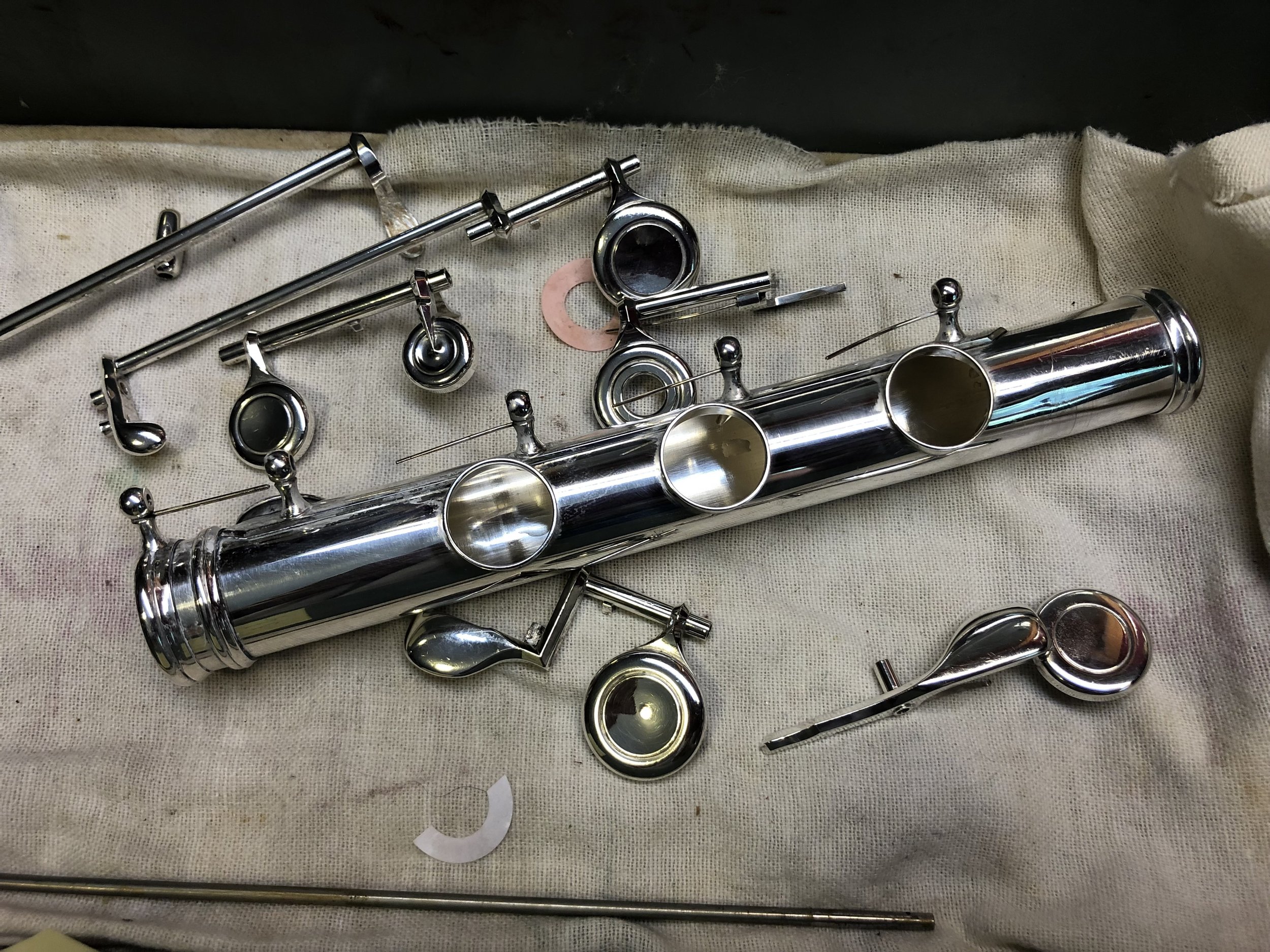 Silver musical instrument parts on a cloth surface, including a flute, tuning keys, rods, and miscellaneous metal pieces.