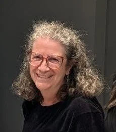 A woman with curly gray hair, glasses, and a black shirt, smiling at the camera.