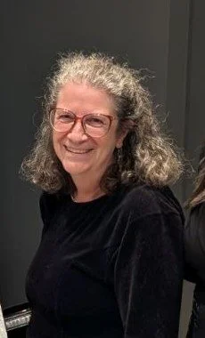 A woman with curly gray hair and glasses smiling, wearing a black top, in an indoor setting.