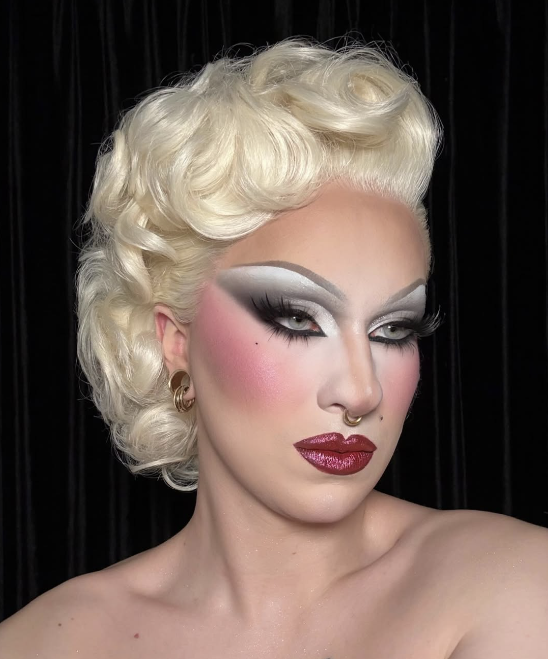 Close-up of a person with platinum blonde, curly hair and glamorous drag makeup, including bold eyebrows, silver and grey eyeshadow, long false eyelashes, pink blush, and dark red lipstick, set against a black background.