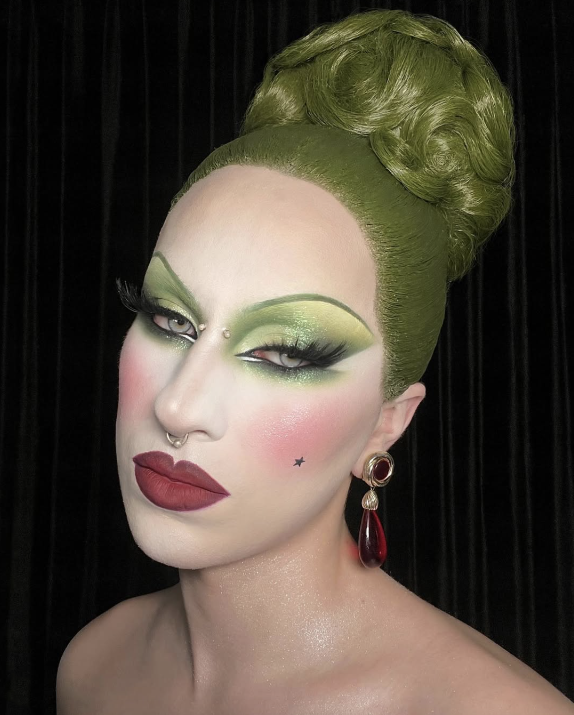 Close-up of a person with elaborate green Marie Antoinette inspired makeup, styled green hair in a beehive, and large red earrings, against a black background.
