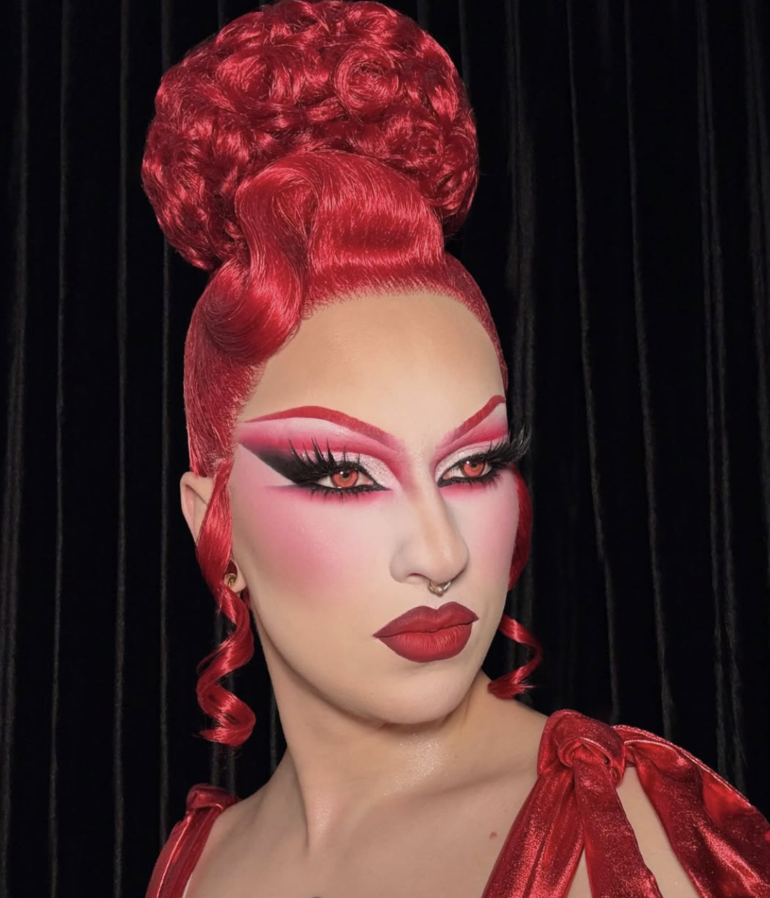 A person with elaborate monochromatic red drag makeup and bright red hair styled in a high, sculpted updo with curls.