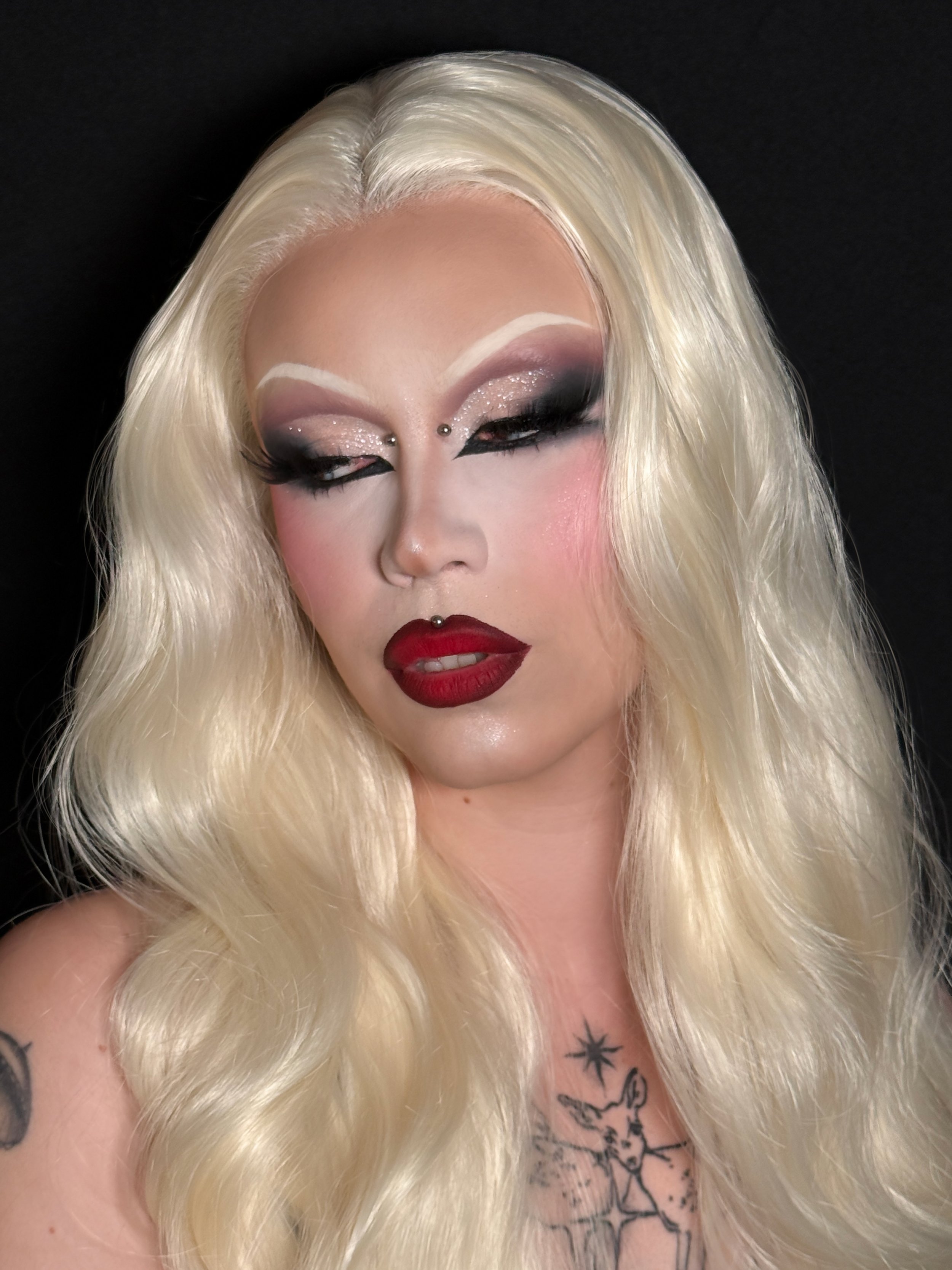 A person with platinum blonde hair and elaborate makeup, including glitter eyeshadow, false eyelashes, and dark lipstick. They have a nose piercing and tattoos on their chest and arm, with a black background.