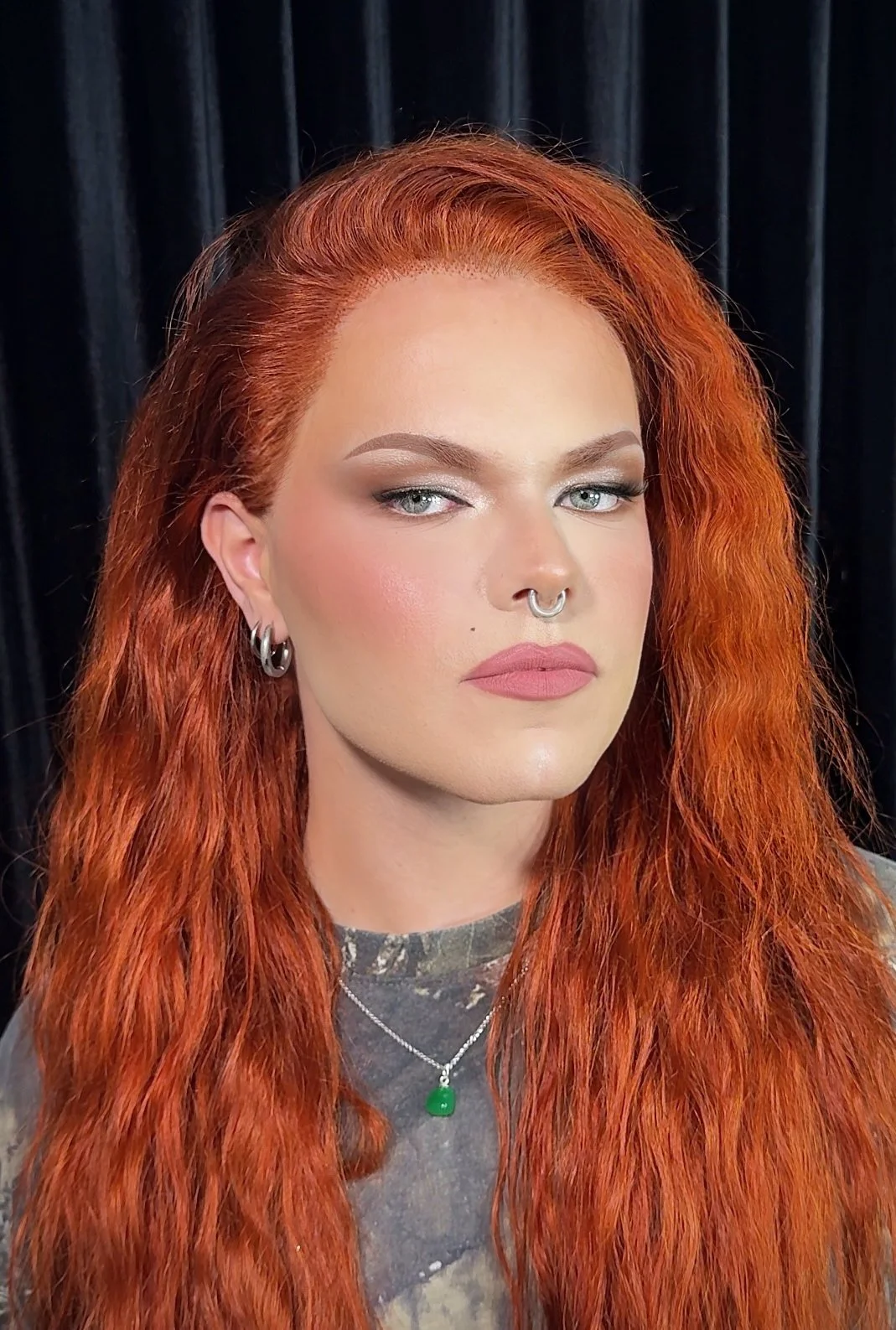 A person with long, wavy red hair, light male to female makeup, and multiple earrings, wearing a silver necklace with a green pendant, standing against a dark background.