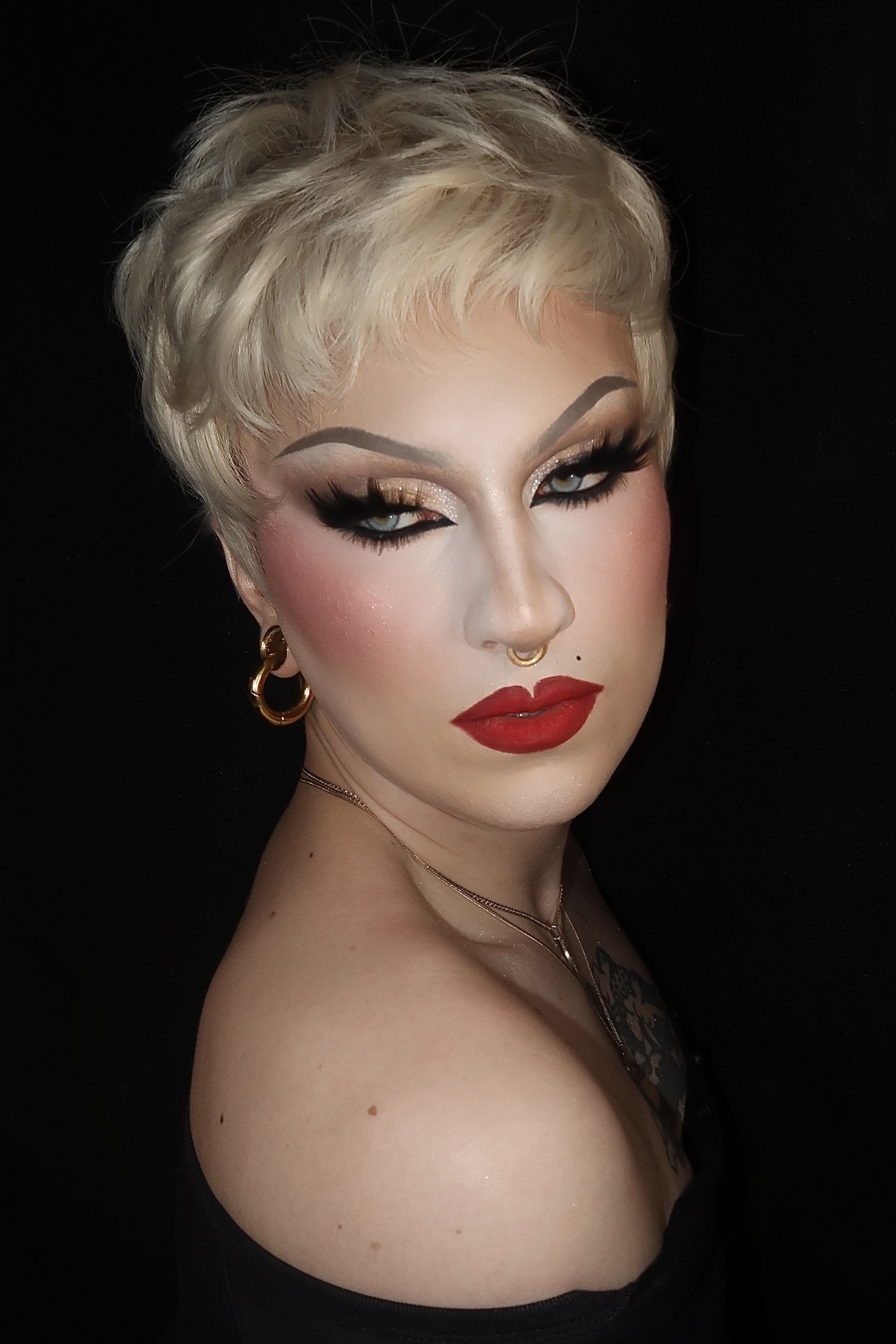 Close-up portrait of a person with platinum blonde hair styled in a short, tousled cut, wearing Madonna inspired drag makeup including bold red lipstick, glitter eyeshadow, and thick black eyeliner, against a black background.