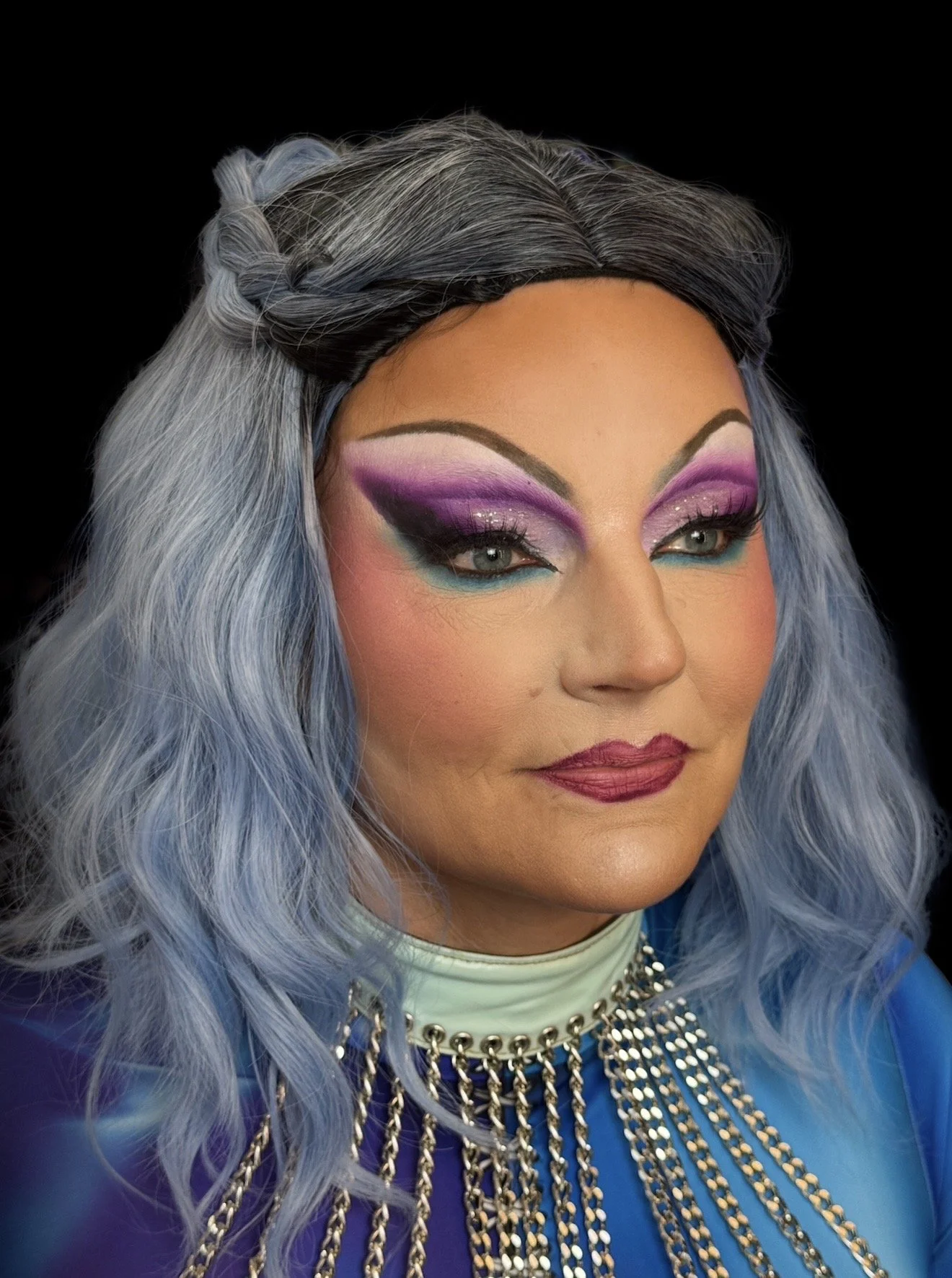 A person in drag makeup with long, wavy blue hair styled with a braid on top. She's wearing vibrant purple eyeshadow with glitter, teal eyeliner, and lipstick. She has a statement necklace with metallic chains, against a black background.