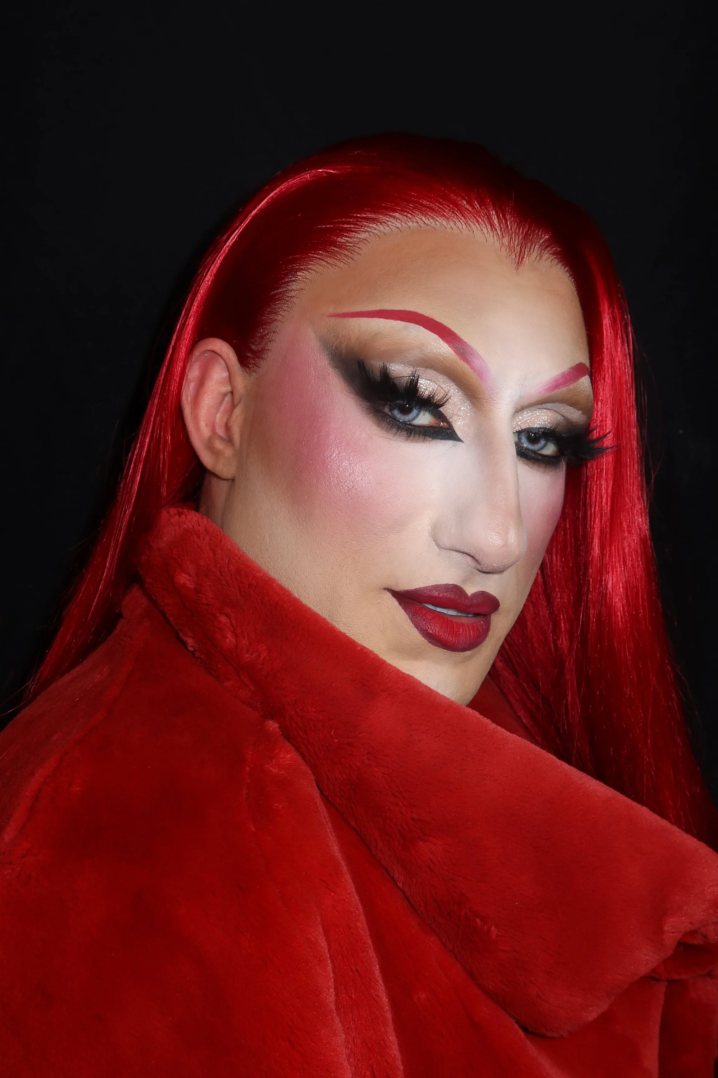 Close-up of a person with bright red hair, bold drag makeup including dark eyeliner, shimmering eyeshadow, pink blush, and deep red lipstick, red eyebrows, wearing a red furry coat against a black background.
