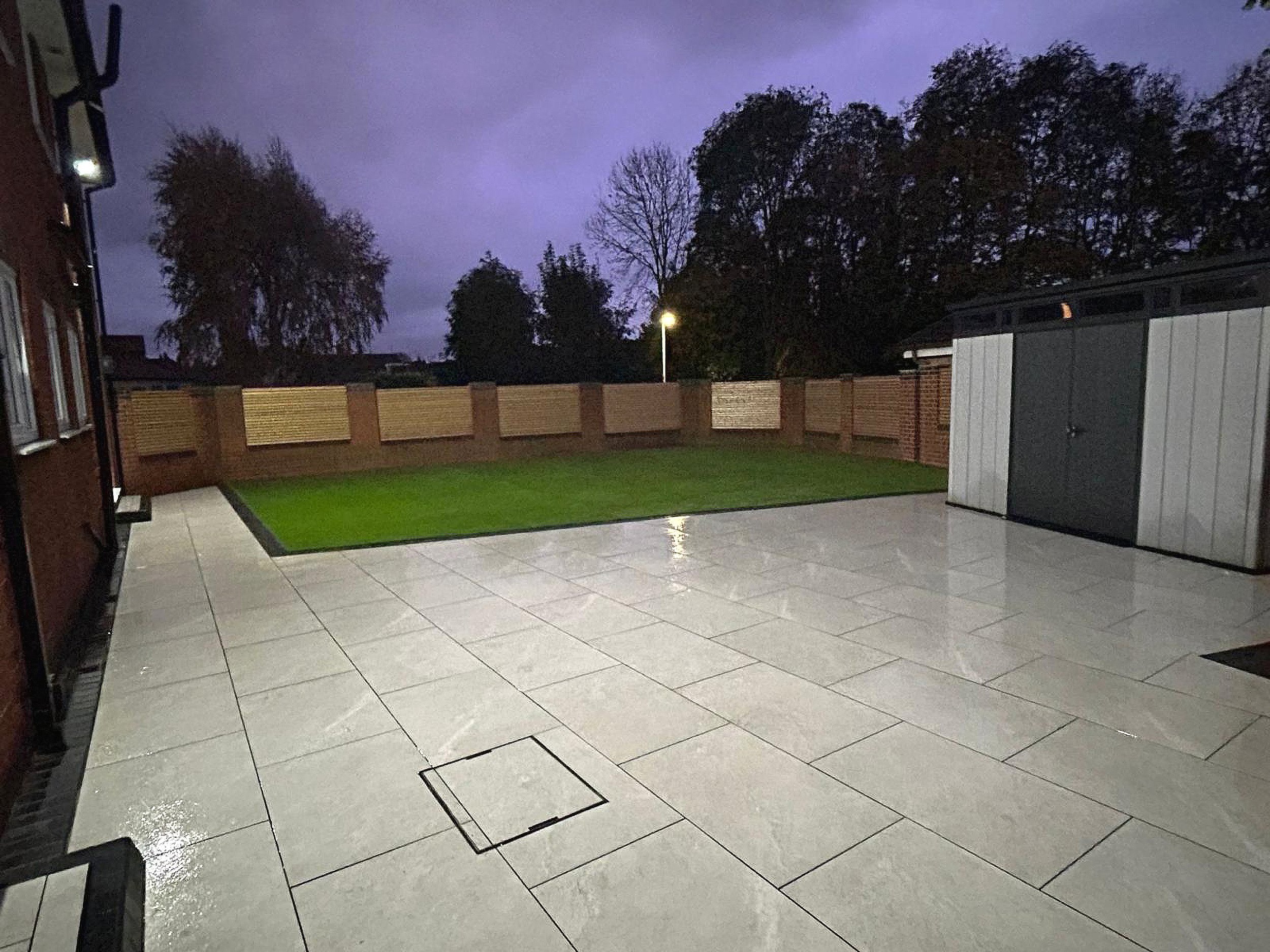 Residential patio with white porcelain tiled flooring, a small patch of artificial grass, a wooden fence, and a shed. There are trees and a streetlamp visible in the background under a dark, cloudy evening sky.