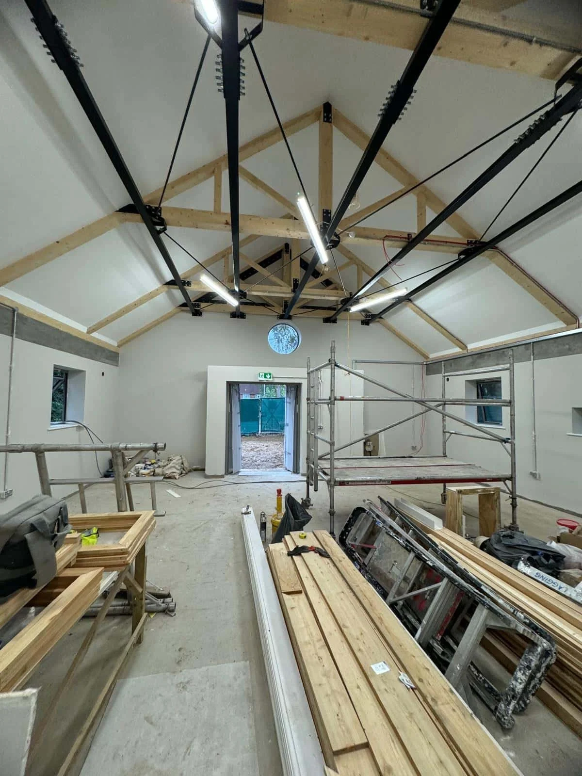 Interior of a building under construction, with scaffolding, construction tools, and framing materials. The space has a high ceiling with exposed wooden beams and lighting.