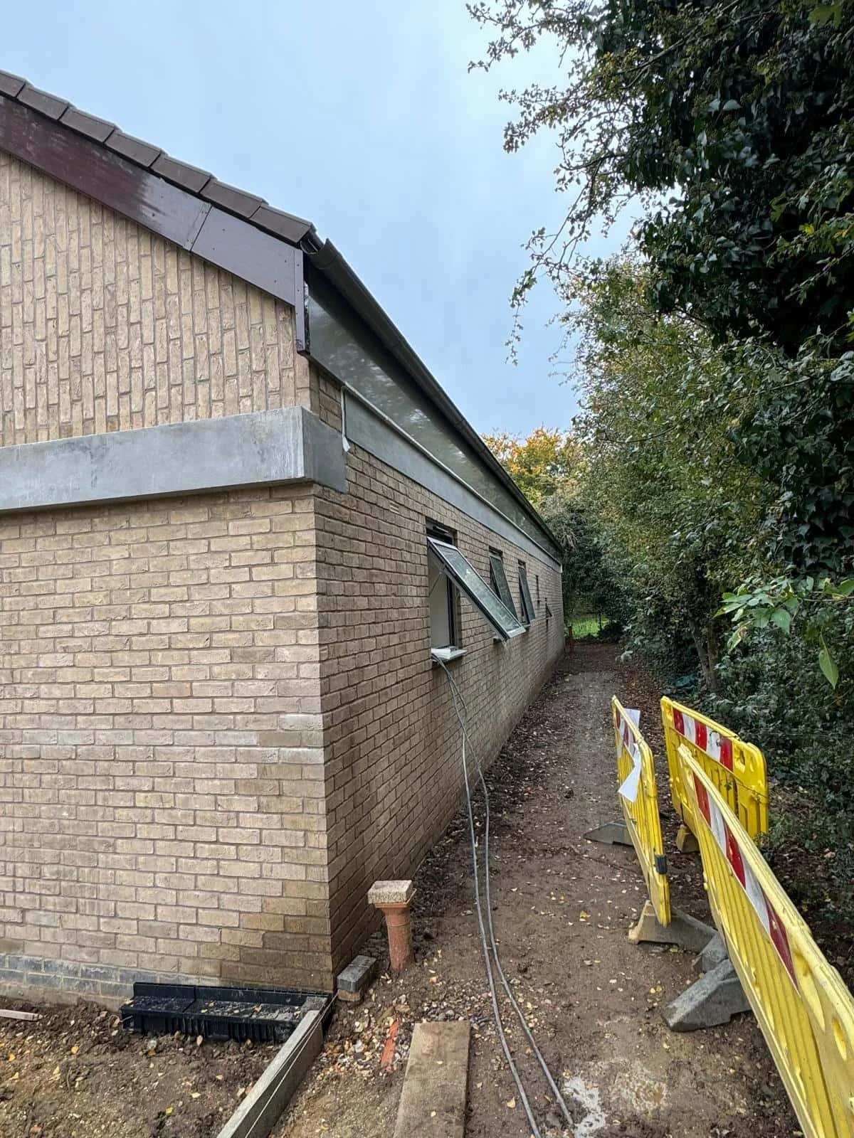 Side view of a brick building under construction with an open window, construction barriers, and construction equipment on a dirt path surrounded by trees.