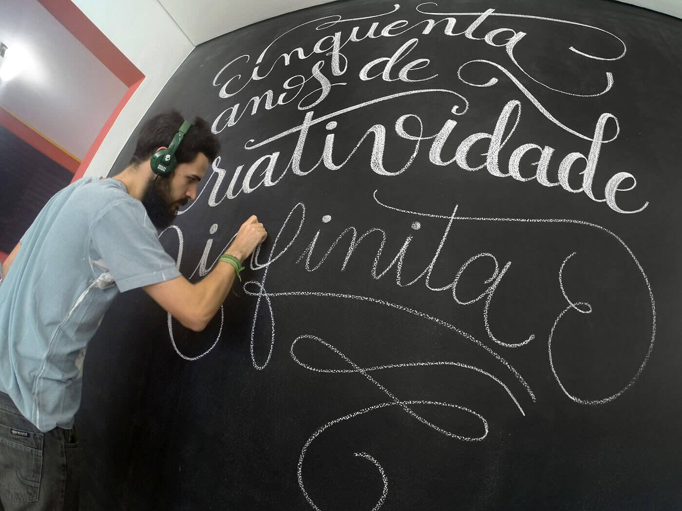 A man with a beard, wearing headphones and a light gray shirt, is writing in white chalk on a large black chalkboard. The chalkboard features cursive text in Spanish that reads 'Chiqueta, amor, de gratitud, agradecimiento, gratitud' with decorative f