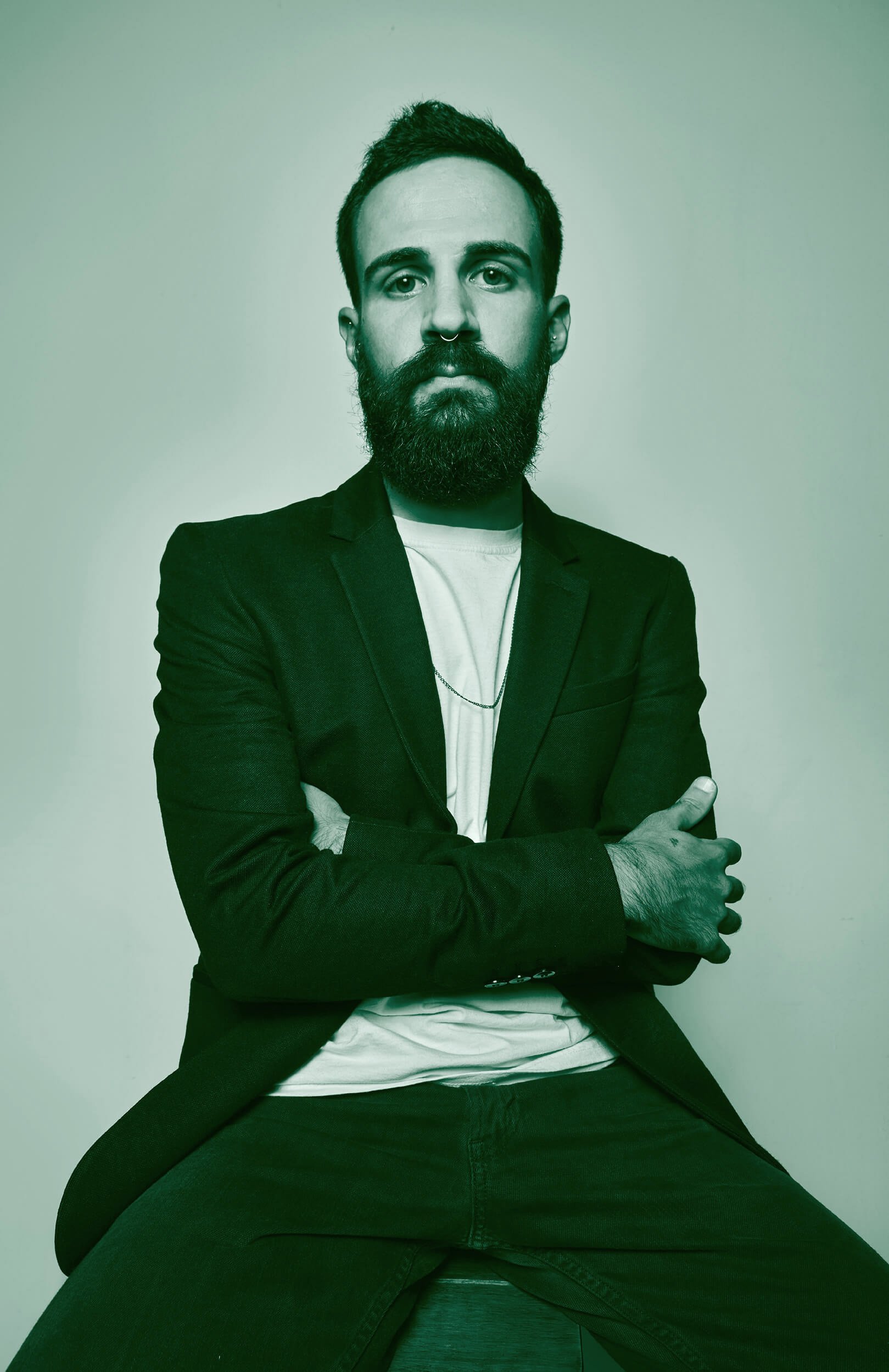 A man with a beard, mustache, and piercings in his nose and ears, wearing a blazer over a white shirt, sitting with his legs spread wide, arms crossed, against a plain background.