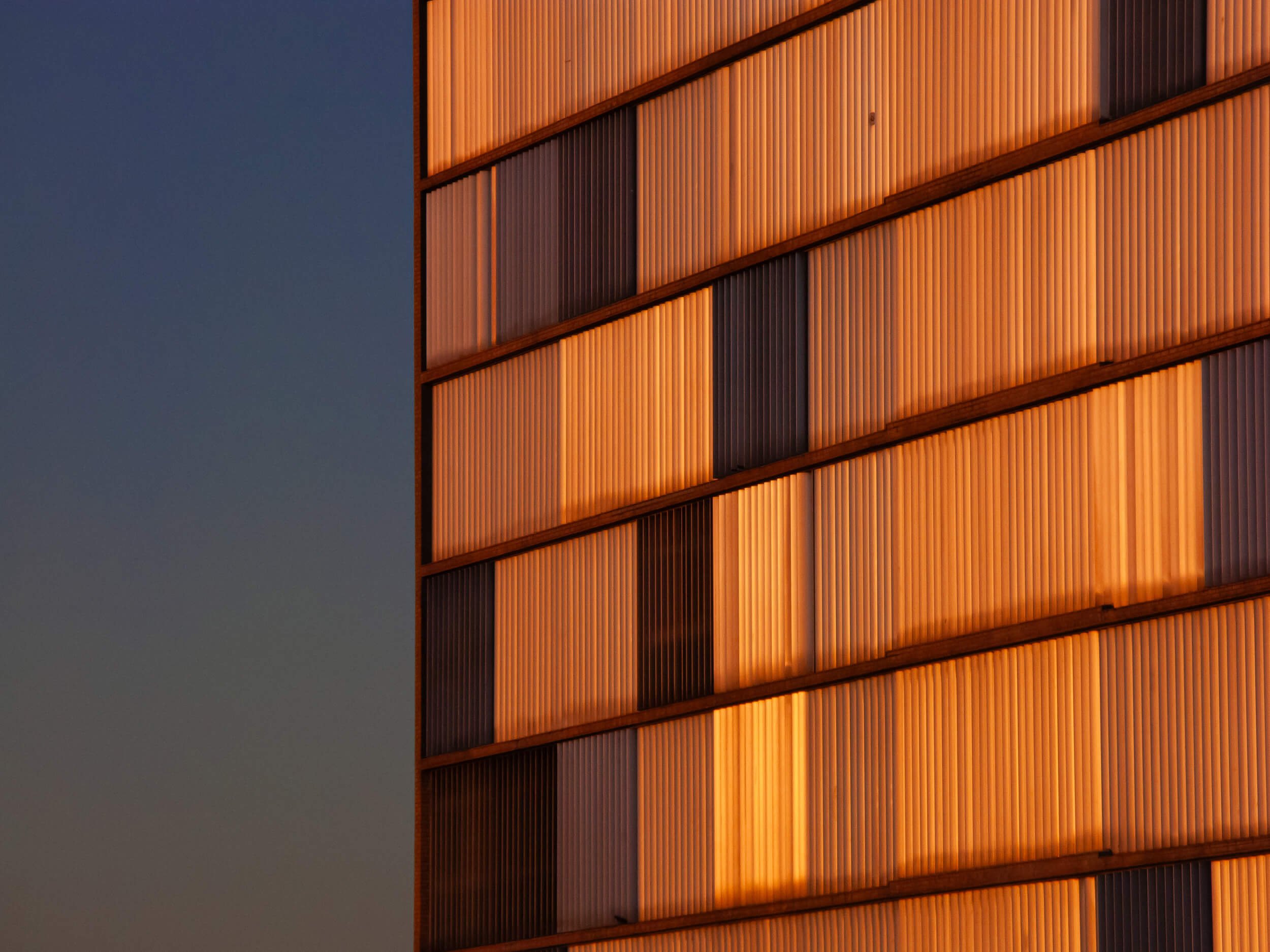 Close-up of a modern building's exterior wall with vertical wooden slats illuminated in warm orange light, set against a dark blue sky.