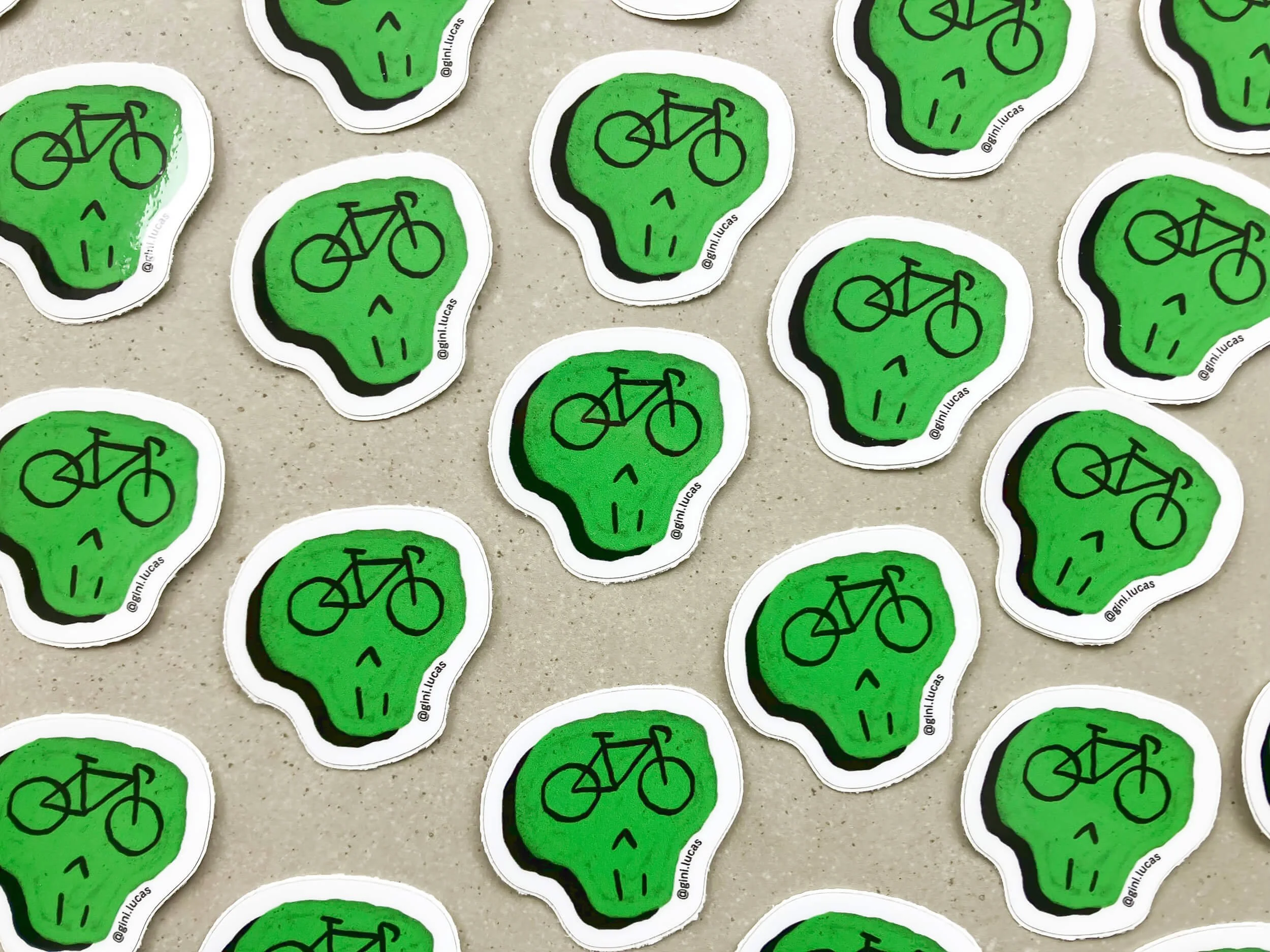 Multiple stickers of a green skull with a bicycle symbol on its forehead, arranged randomly on a beige surface.