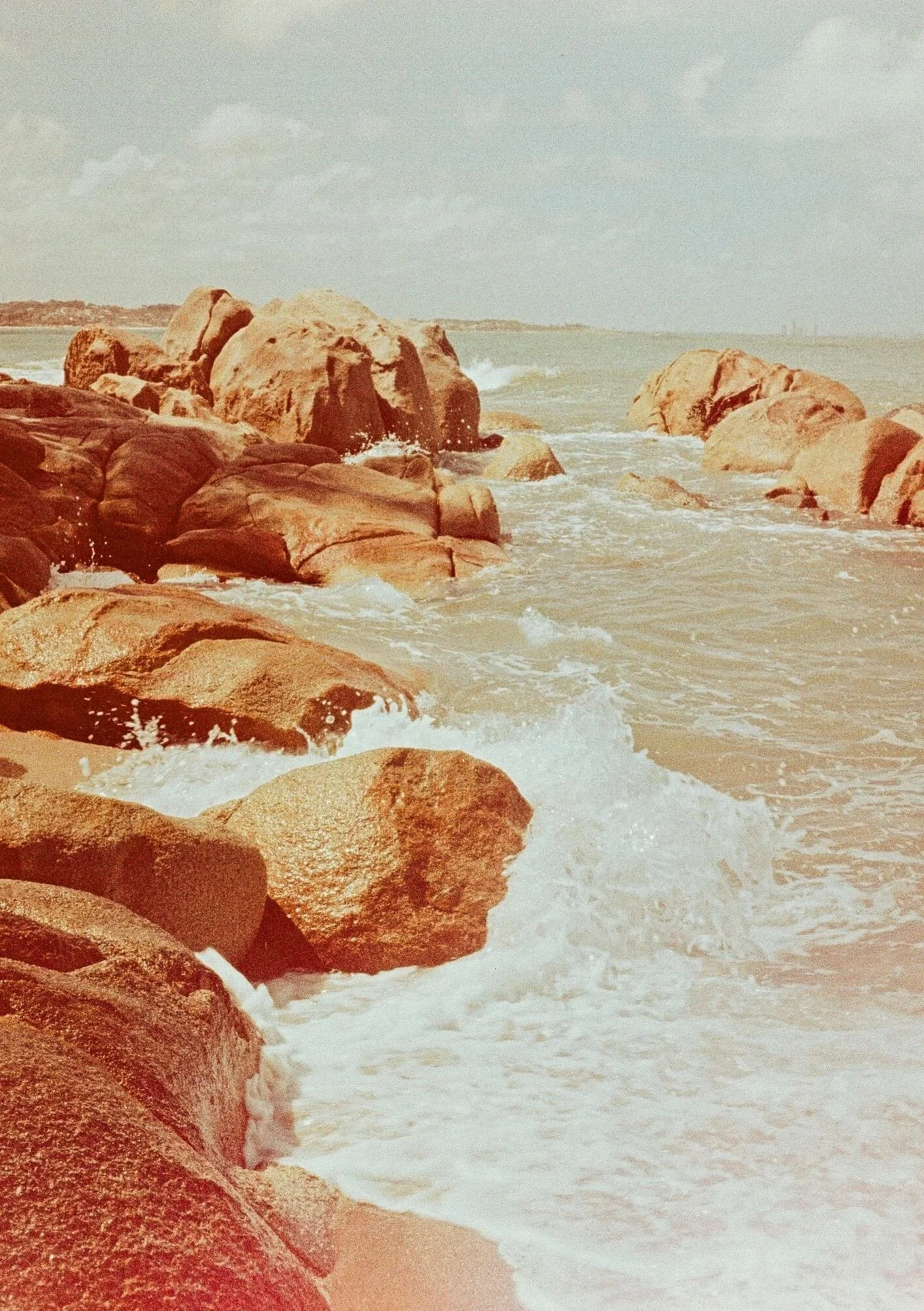 Large rocks along a shoreline with ocean waves crashing against them, distant land in the background under a partly cloudy sky.