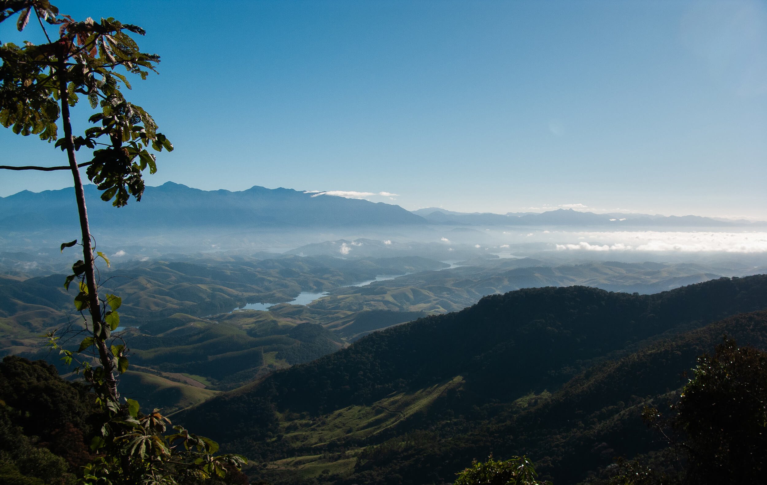 Scenic view of rolling green hills and valleys with a river winding through, mountain range in the background, and a clear blue sky with some clouds.