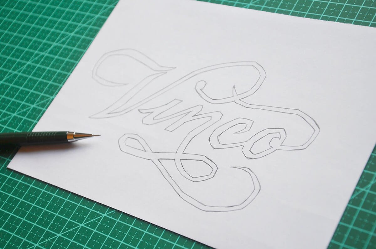 Outline sketch of the word "Vinco" on a white sheet of paper, placed on a green cutting mat, with a black pen lying next to it.